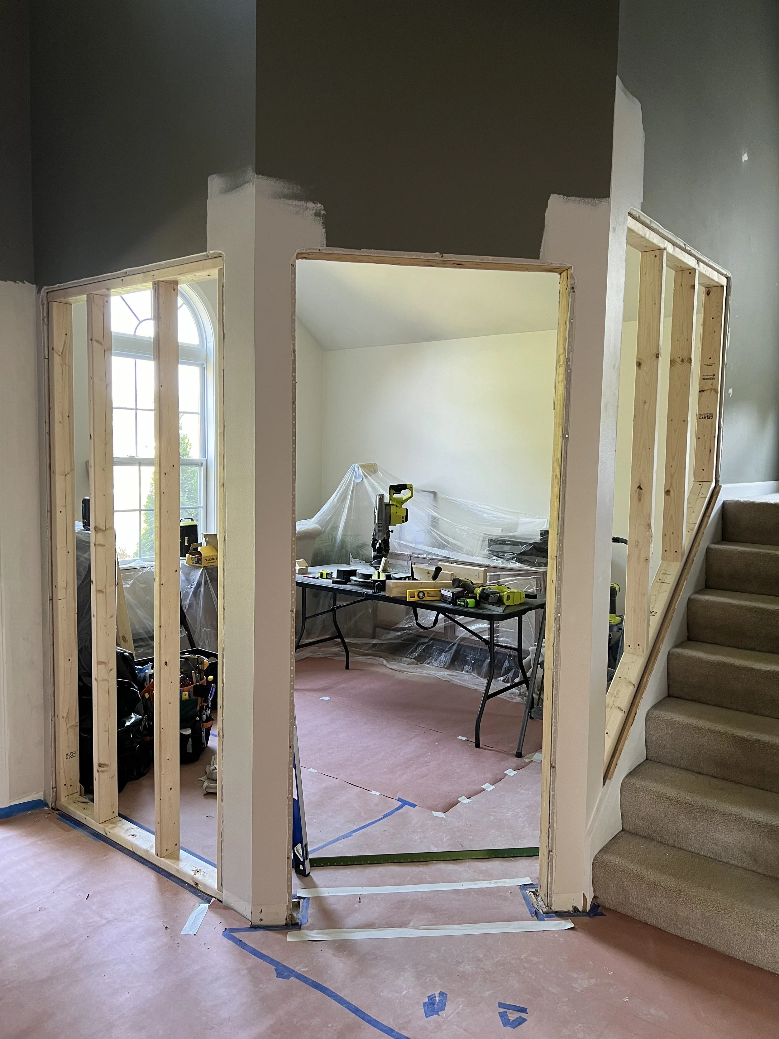 Interior remodeling of a home with new wall framing and construction tools, including a drill and a saw, around an archway near a staircase, with people working behind plastic sheeting in the background.