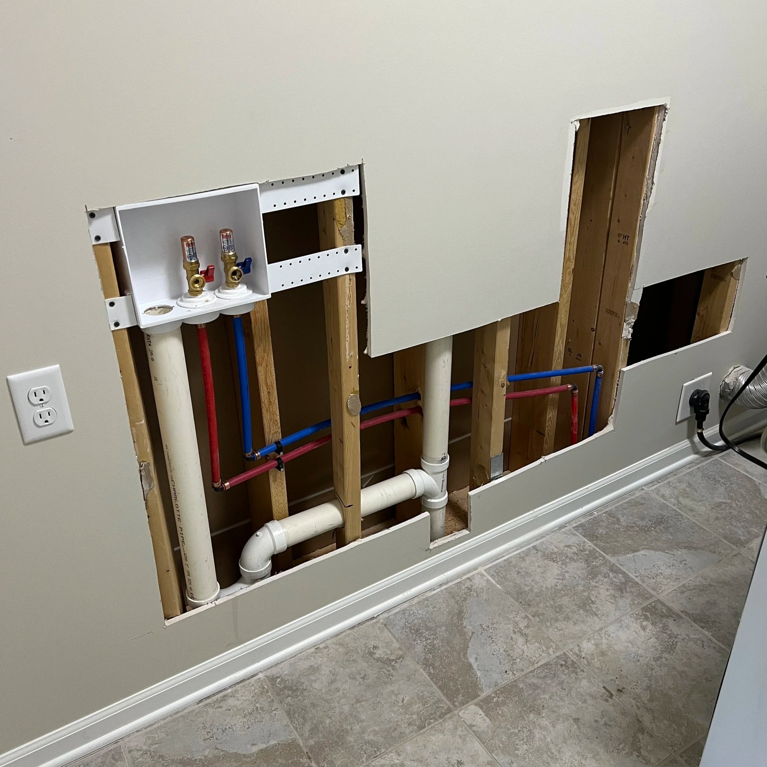 The image shows a wall with plumbing and pipes for a future laundry or utility installation, with two holes cut in the drywall for access.