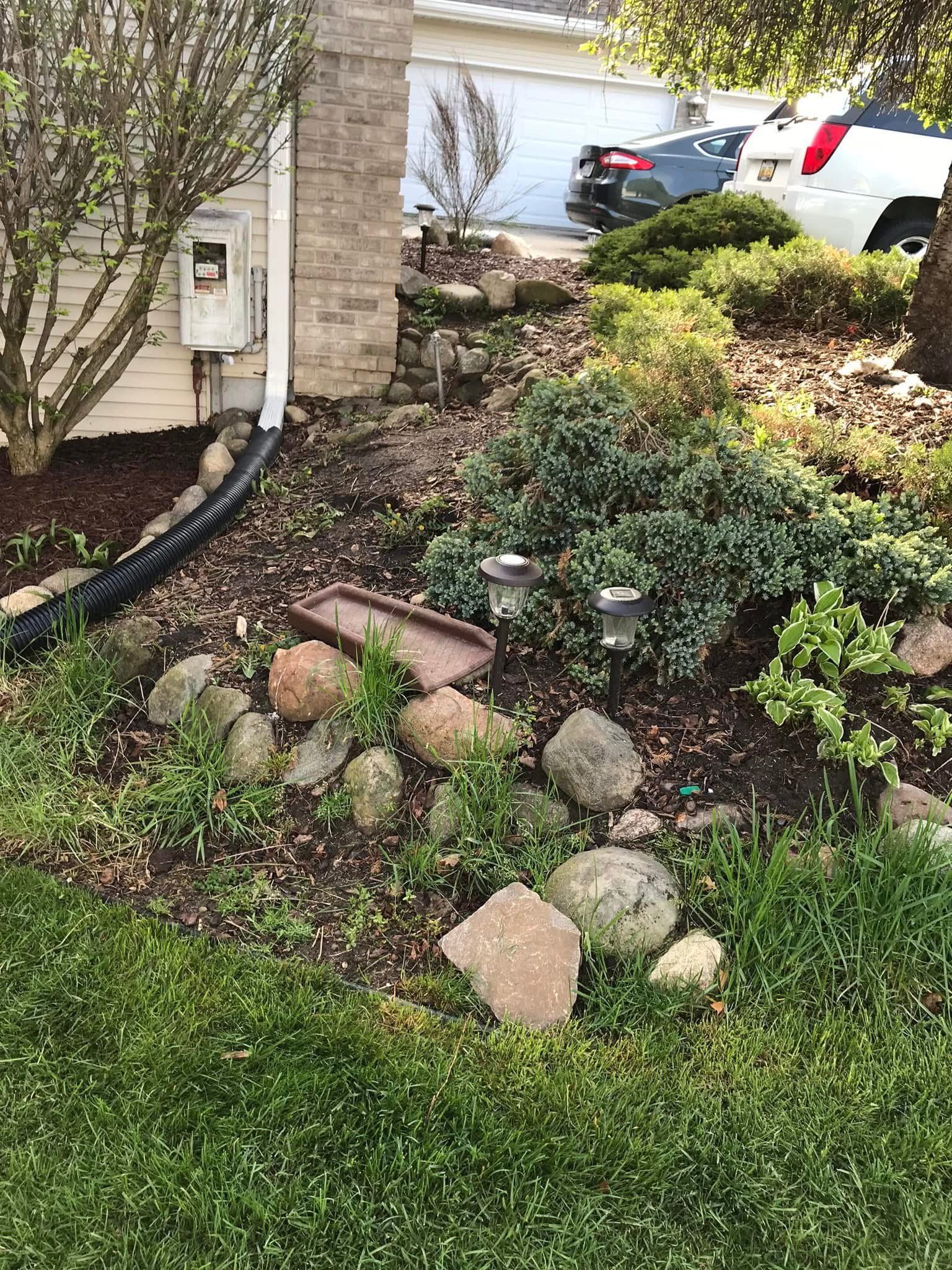 A landscaped front yard with a garden bed, rocks, plants, and garden lights, near a house with a brick wall and a driveway with parked cars.