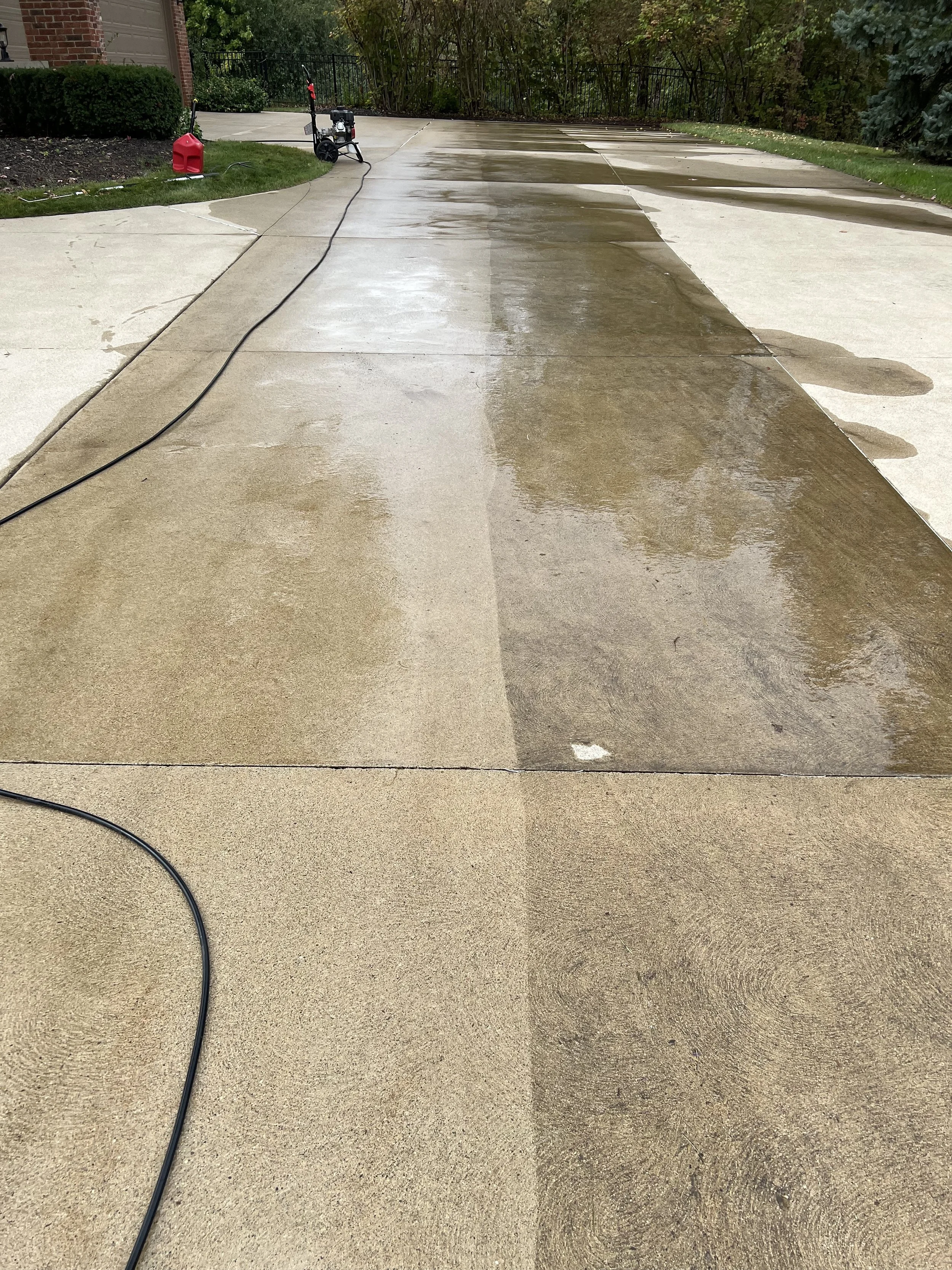 Concrete driveway being cleaned and rinsed with a pressure washer, wet sections visible with water reflecting the surrounding trees.