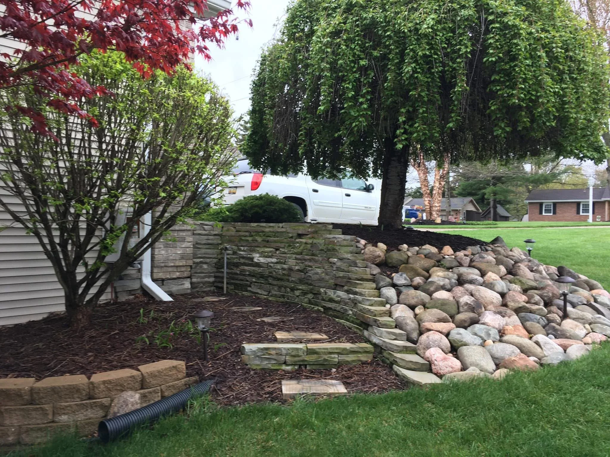 A landscaped yard with a stone retaining wall and a large tree, nearby shrubs, grass, and outdoor lighting