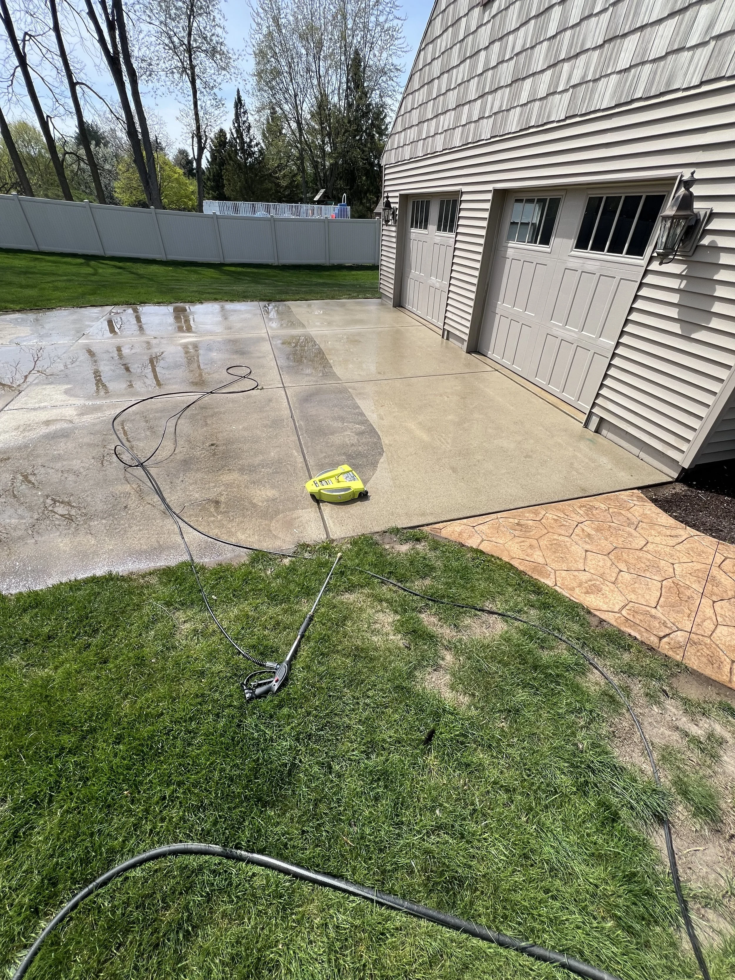 A residential backyard with a concrete patio and a grassy area. A lawn sprinkler is placed on the grass, and garden hoses are spread across the area. The house has beige siding, a garage with two doors, and a lantern-style light fixture. The sky is clear, and trees are visible in the background.