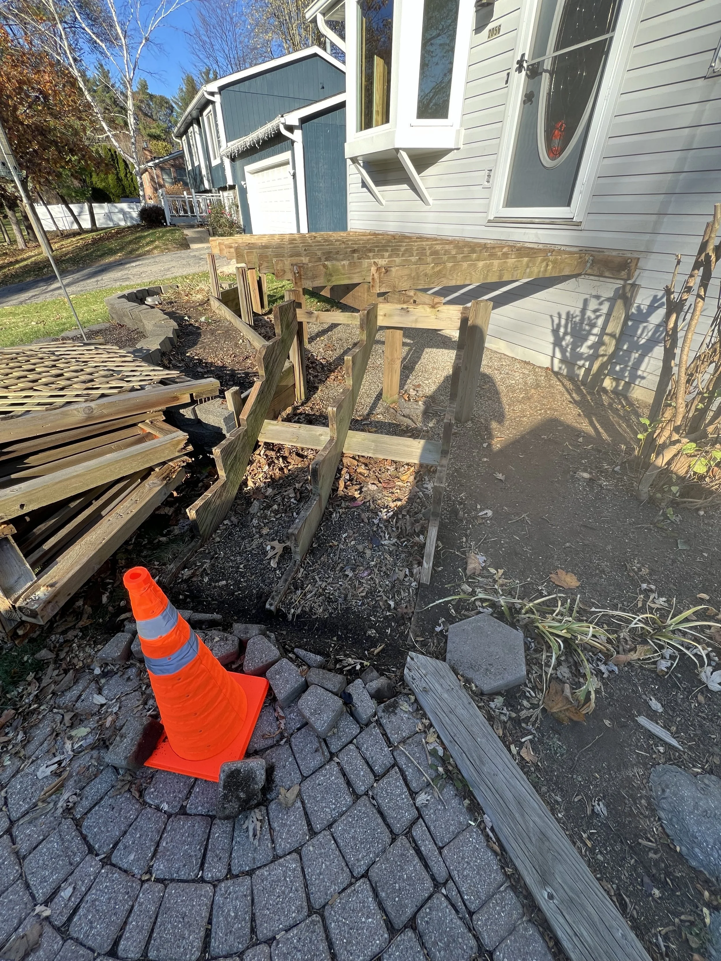 Construction site with a partially built wooden deck attached to a light-colored house, an orange safety cone, and a stack of paving stones, with a house and trees in the background.
