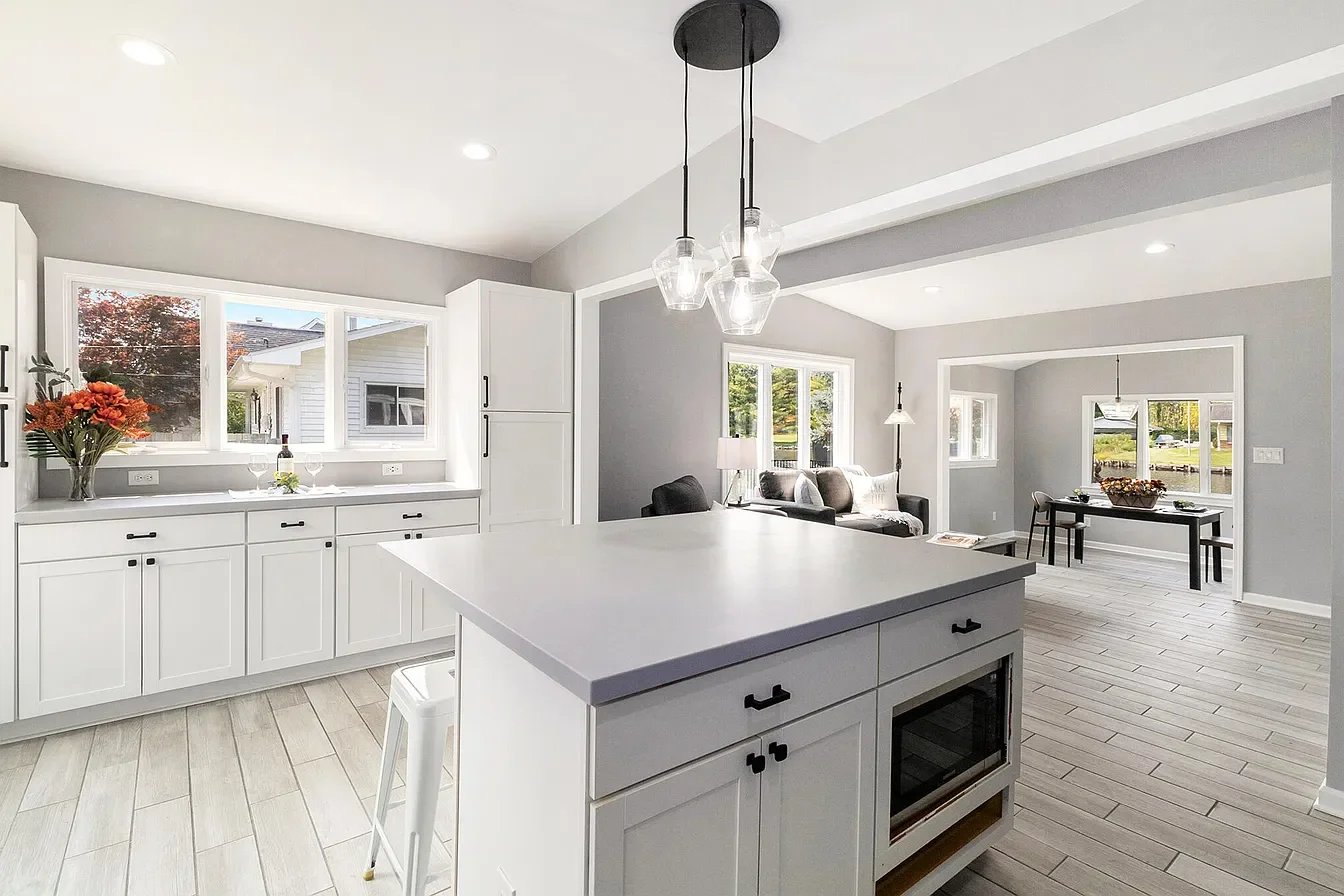 Bright, modern open-concept kitchen and living room with white cabinetry, light gray walls, large windows, and minimal decor. Includes a kitchen island with a built-in microwave, hanging pendant lights, and a sitting area with a sofa and dining table.