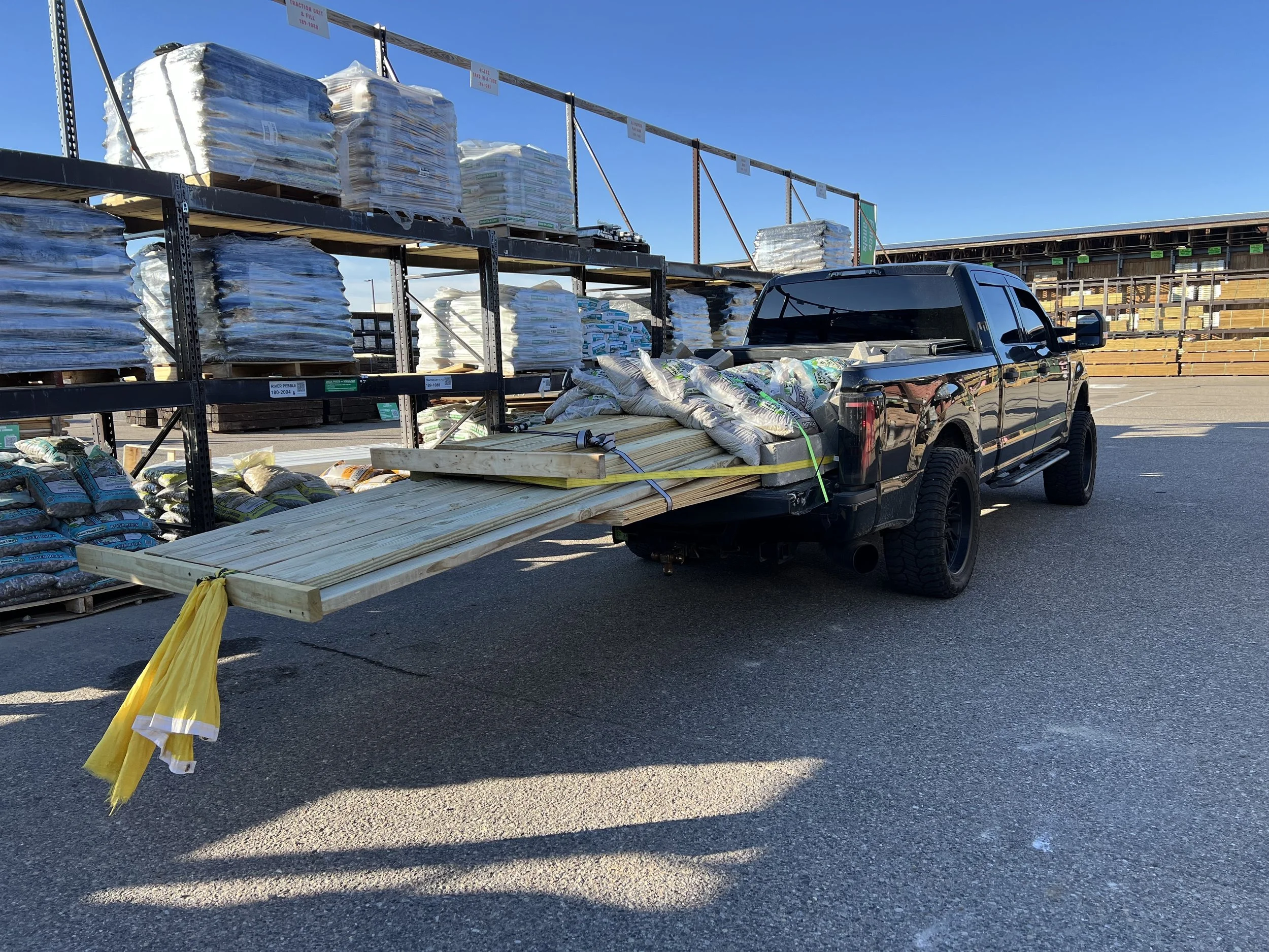 A black pickup truck loaded with wooden planks and bags of materials, parked next to metal and wooden shelves filled with other construction supplies at an outdoor store or warehouse.