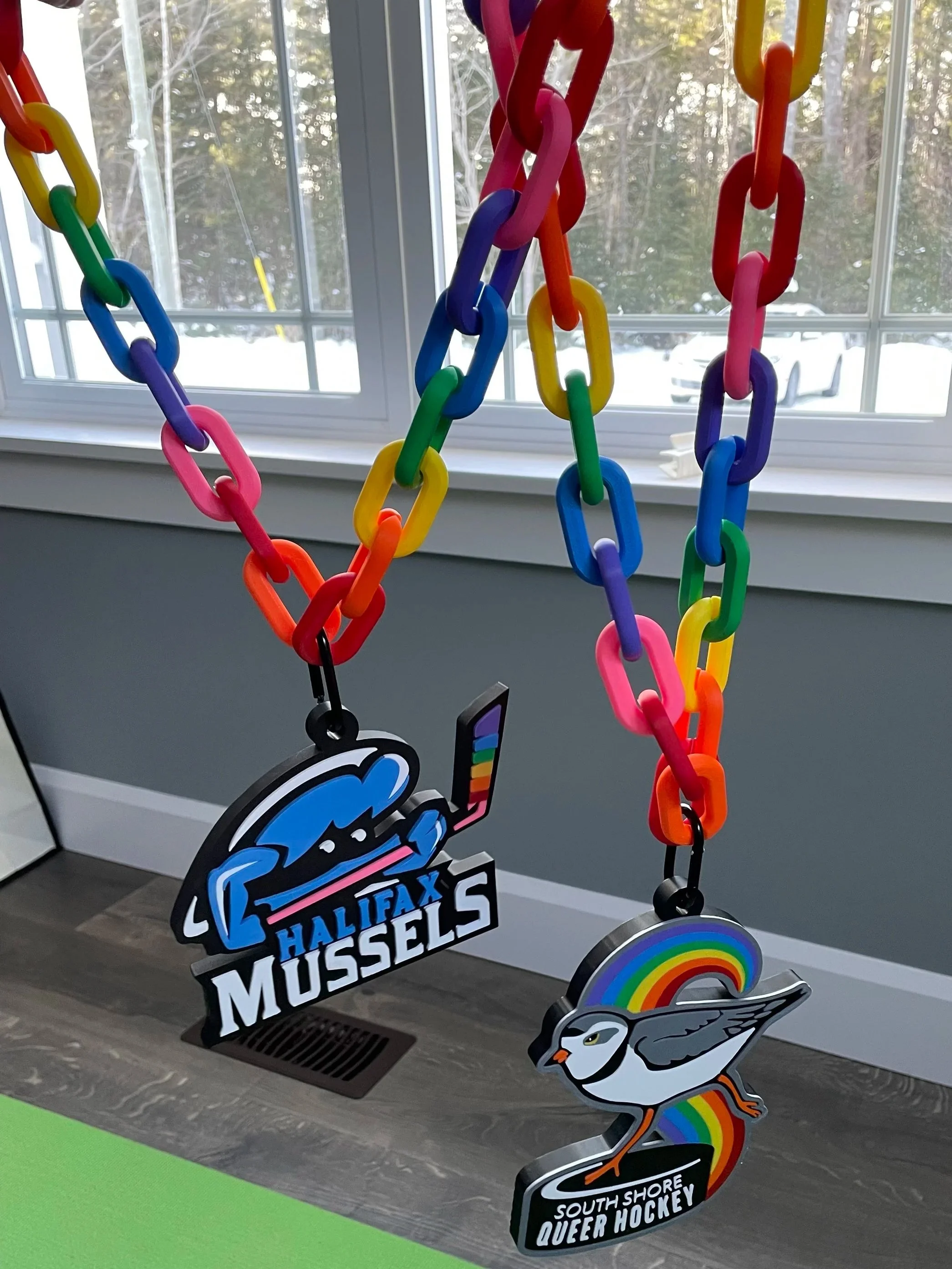 Colorful rainbow plastic chains hanging with two keychains; one features the logo of Halifax Mussels, and the other has a rainbow and bird illustration for South Shore Queer Hockey, near a window with trees outside.