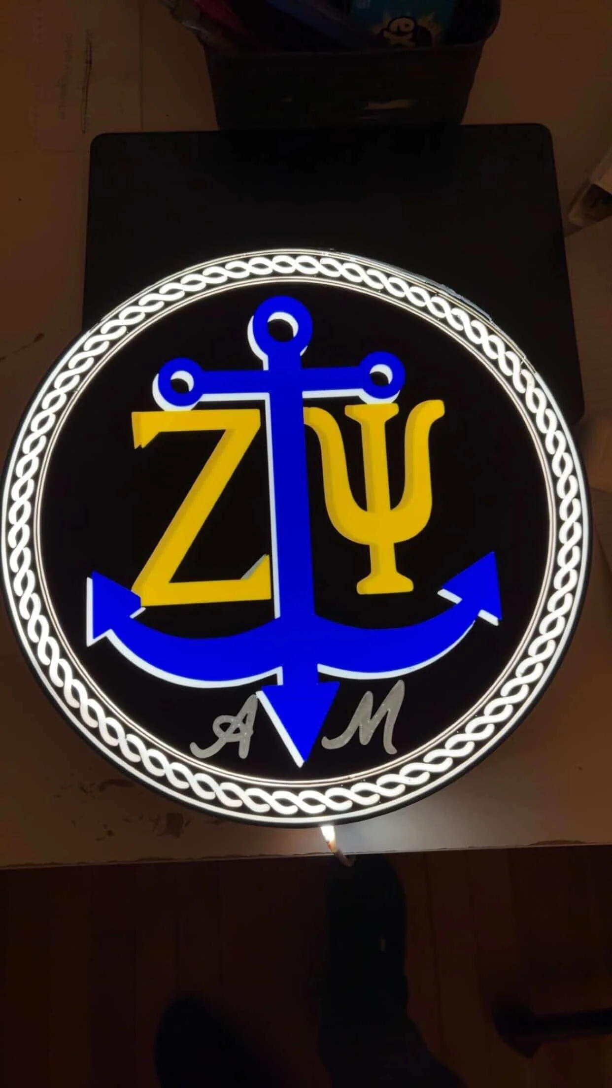 Illuminated sign with a blue anchor, a yellow Greek letter Zeta, a yellow Greek letter Psi, and the letters A and M at the bottom, bordered by a white chain design.