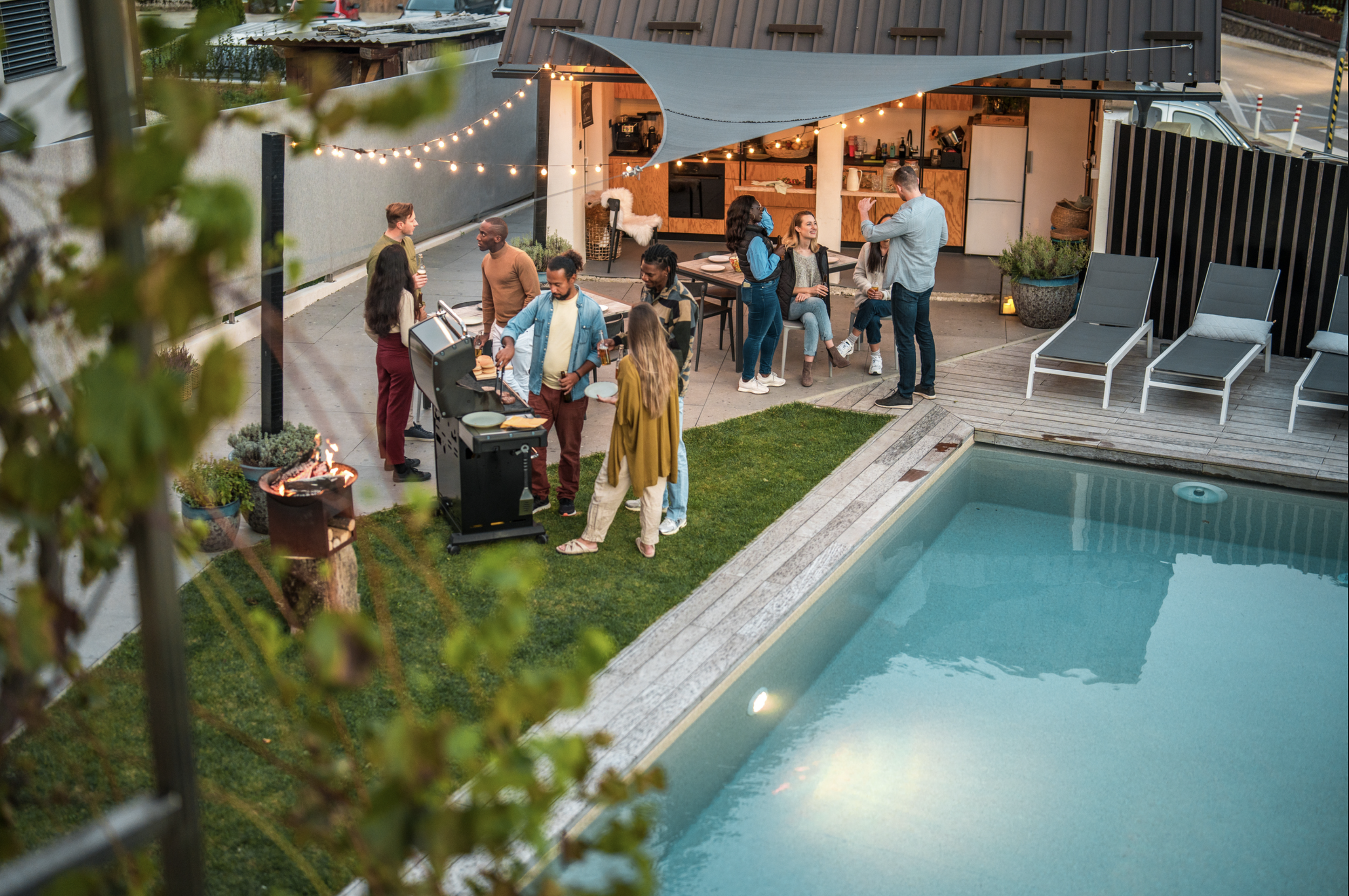 People gathered around a barbecue grill and sitting on a patio near a pool at an outdoor party in the evening, with string lights overhead.