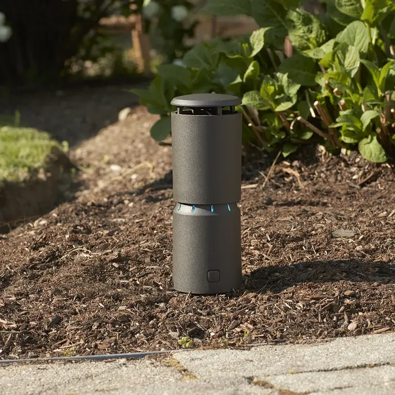 A brown Thermacell LIV mosquito repeller installed in a garden with green plants in the background.