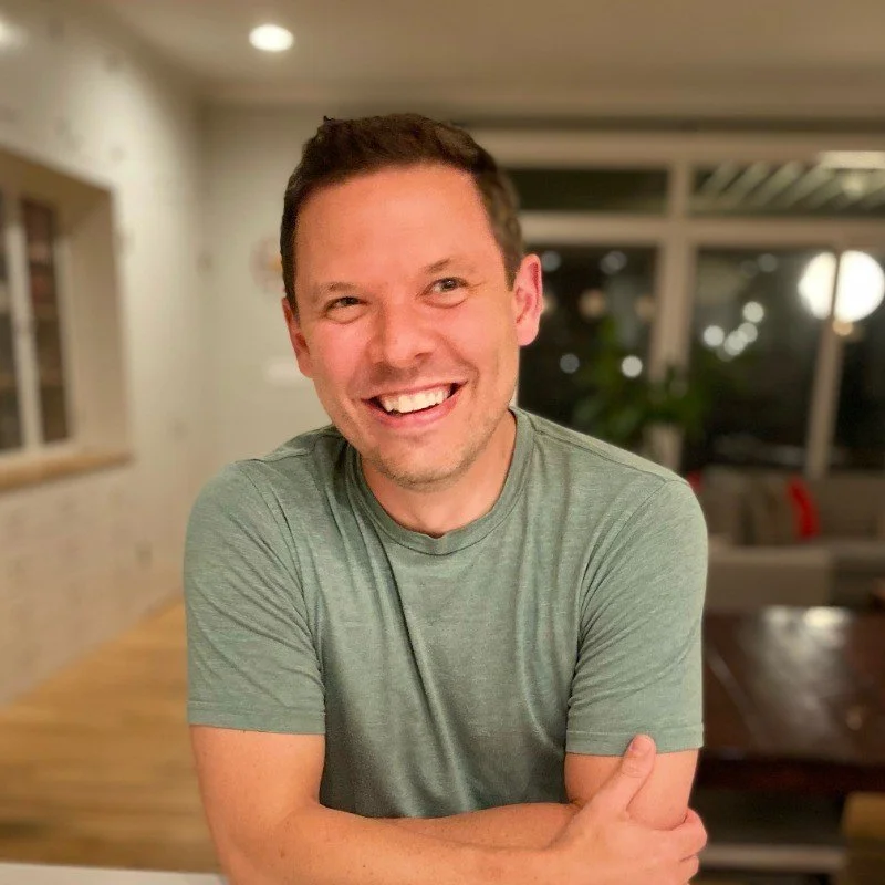 A smiling man with short dark hair and a beard, wearing a green T-shirt, sitting at a table in a well-lit modern room with large windows and a blurred background.