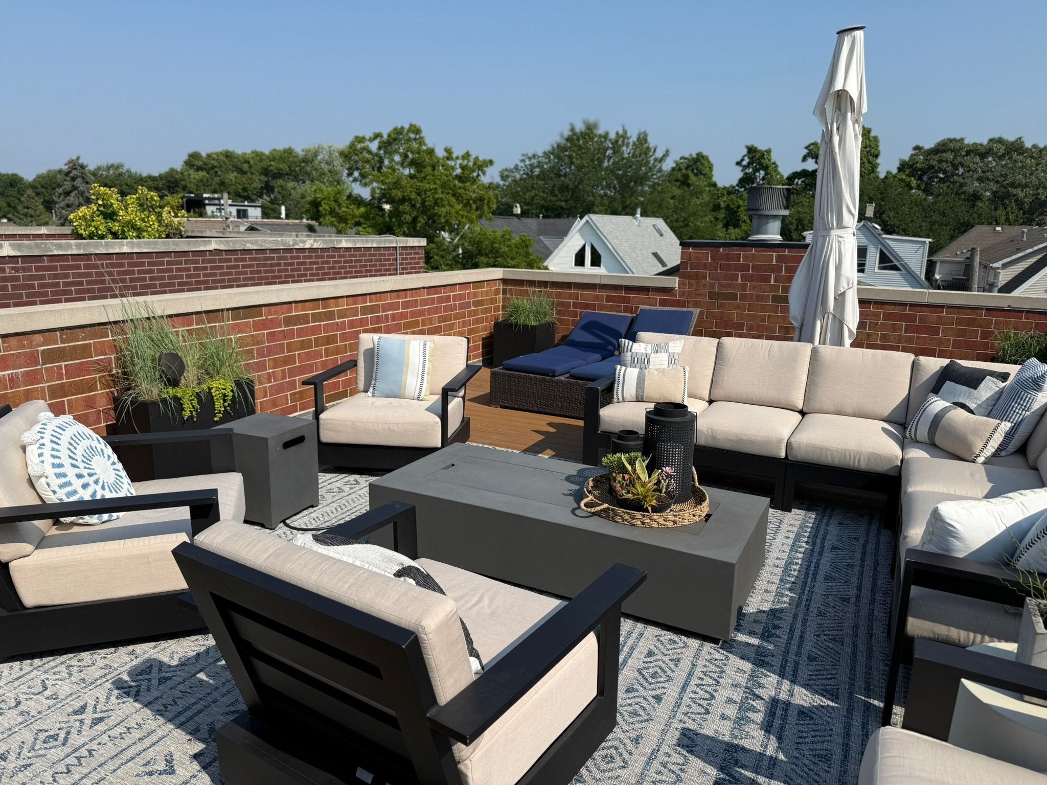 Outdoor rooftop patio with beige cushioned furniture, black tables, and potted plants, enclosed by brick walls, under a clear blue sky.