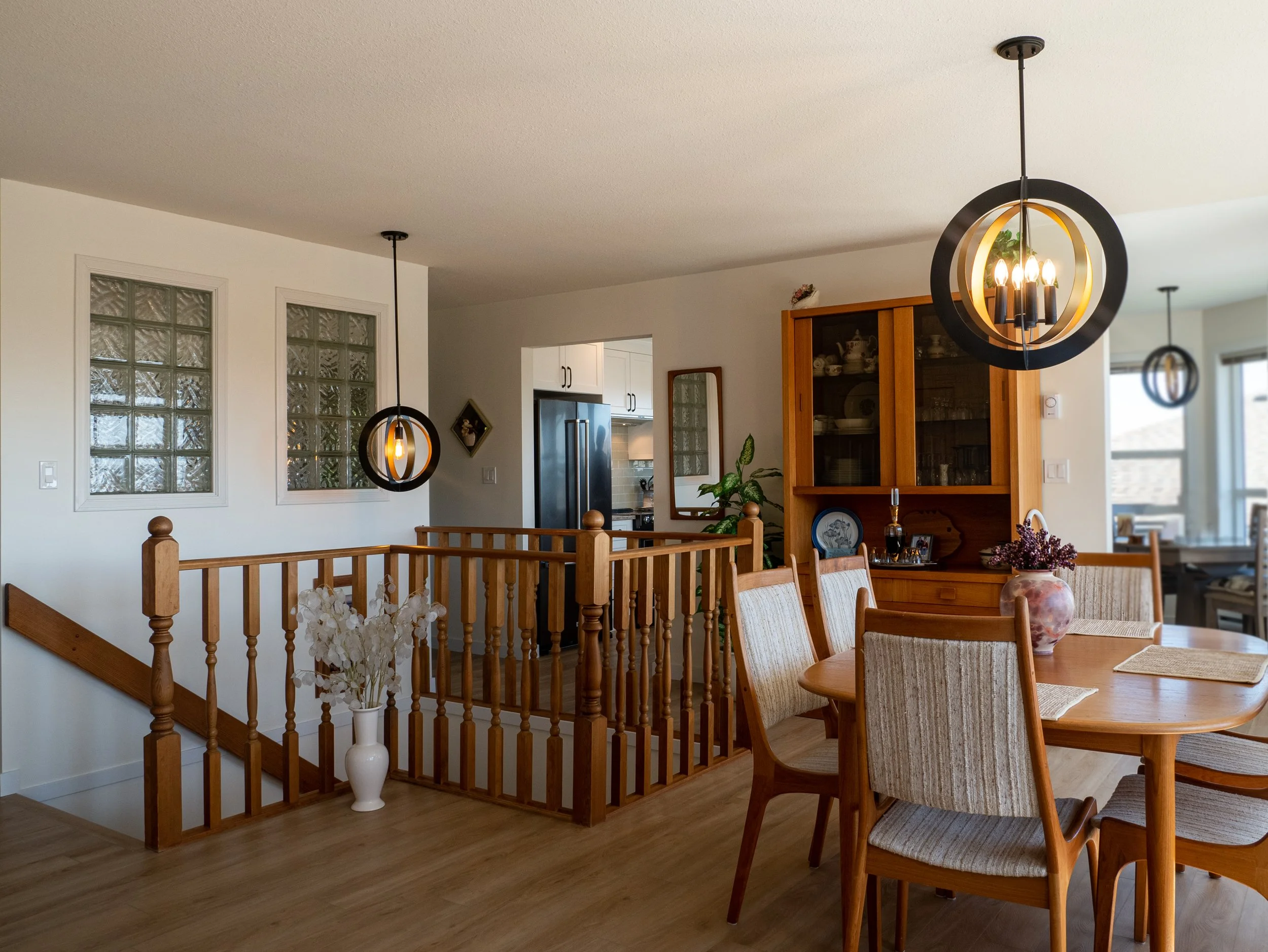 A cozy living space with wooden furniture, featuring a dining table with chairs, a display cabinet, and decorative items including a vase with flowers. Modern hanging light fixtures illuminate the room, which has wooden flooring and white walls with glass block windows.