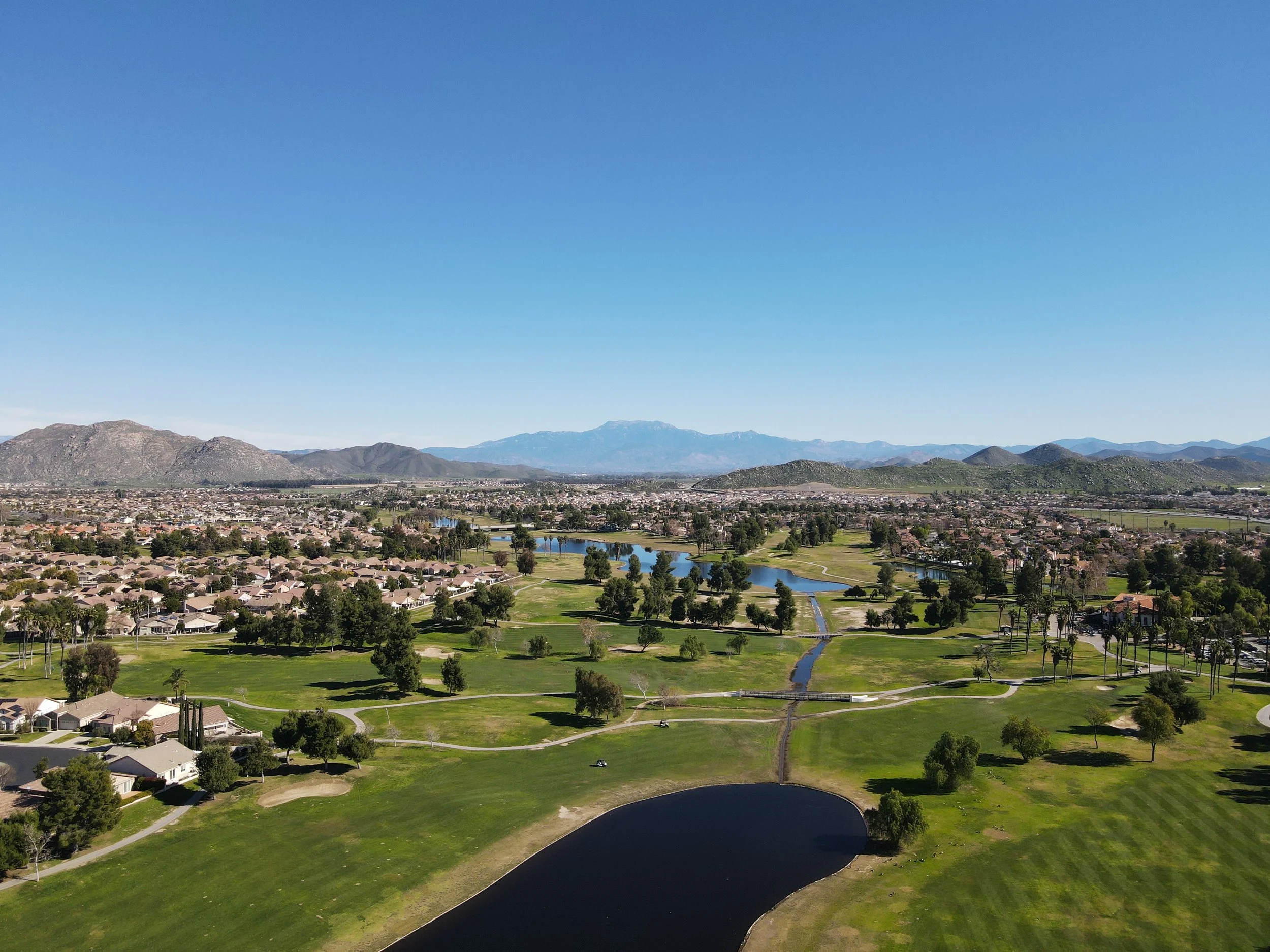 Aerial view of a golf course with water features, surrounded by residential houses and a cityscape with mountains in the background under a clear blue sky.