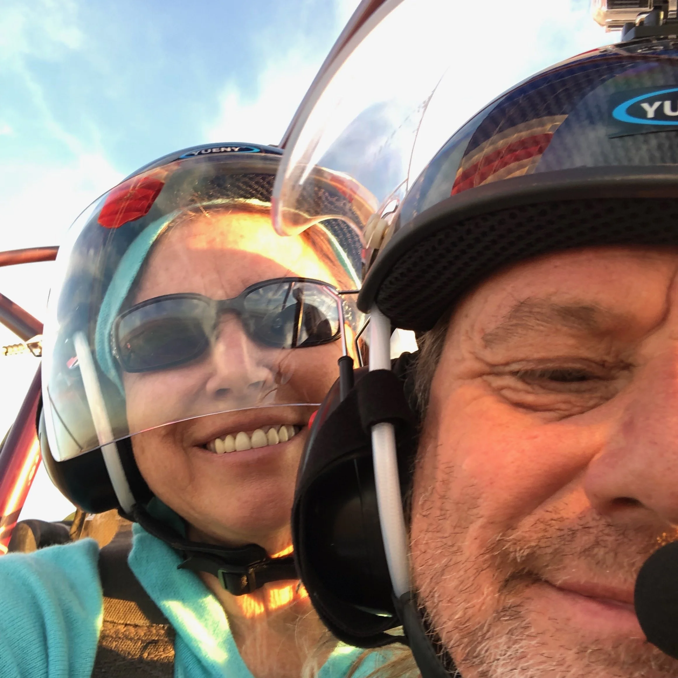 Close-up of a smiling woman and a man in helmets with microphones, appearing to be in a vehicle or aircraft, with a blue sky background.