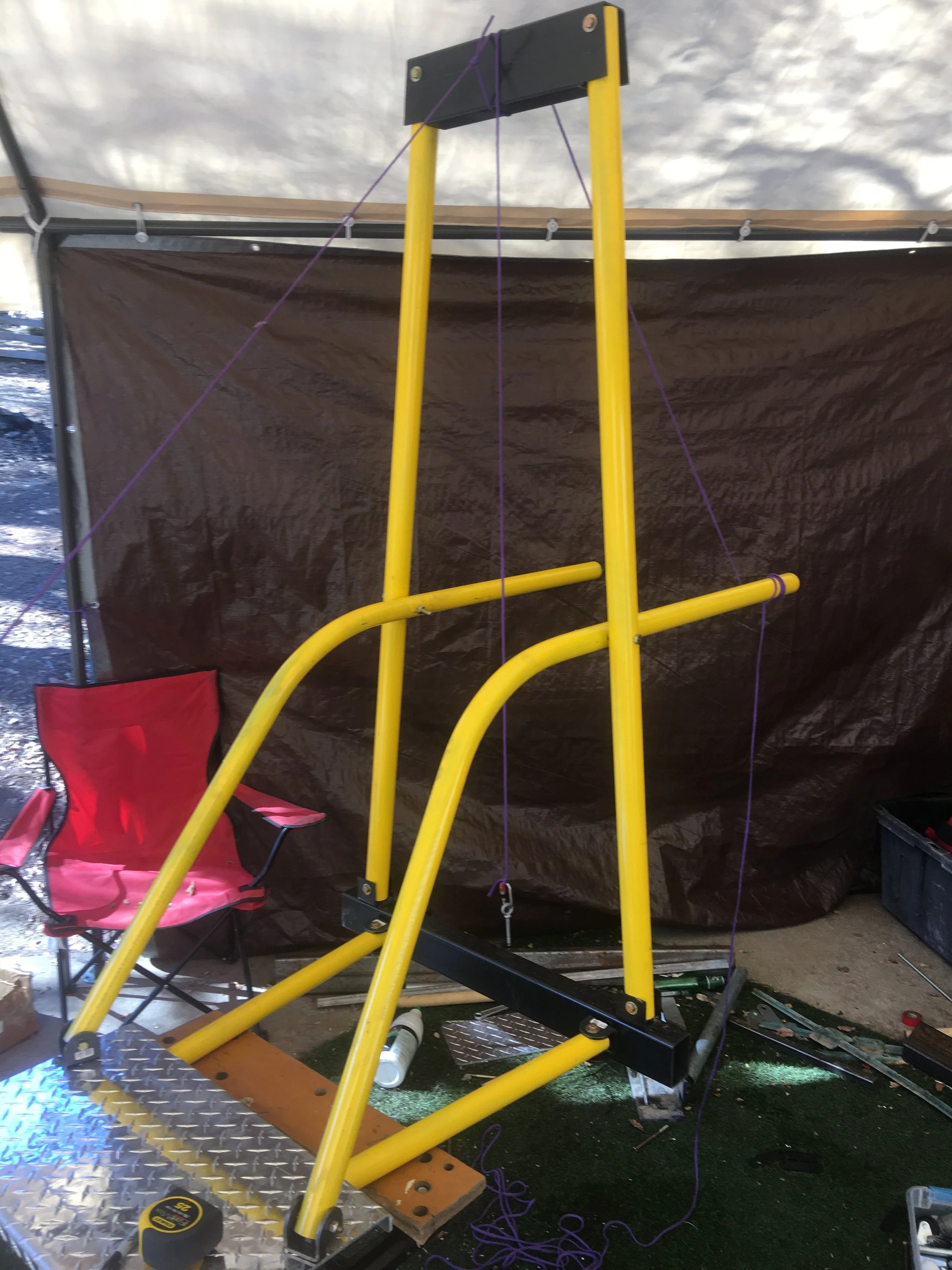 Yellow metal A-frame structure with purple strings attached, set up on a workshop table, with a red folding chair and black storage container nearby, in an indoor or tented area.