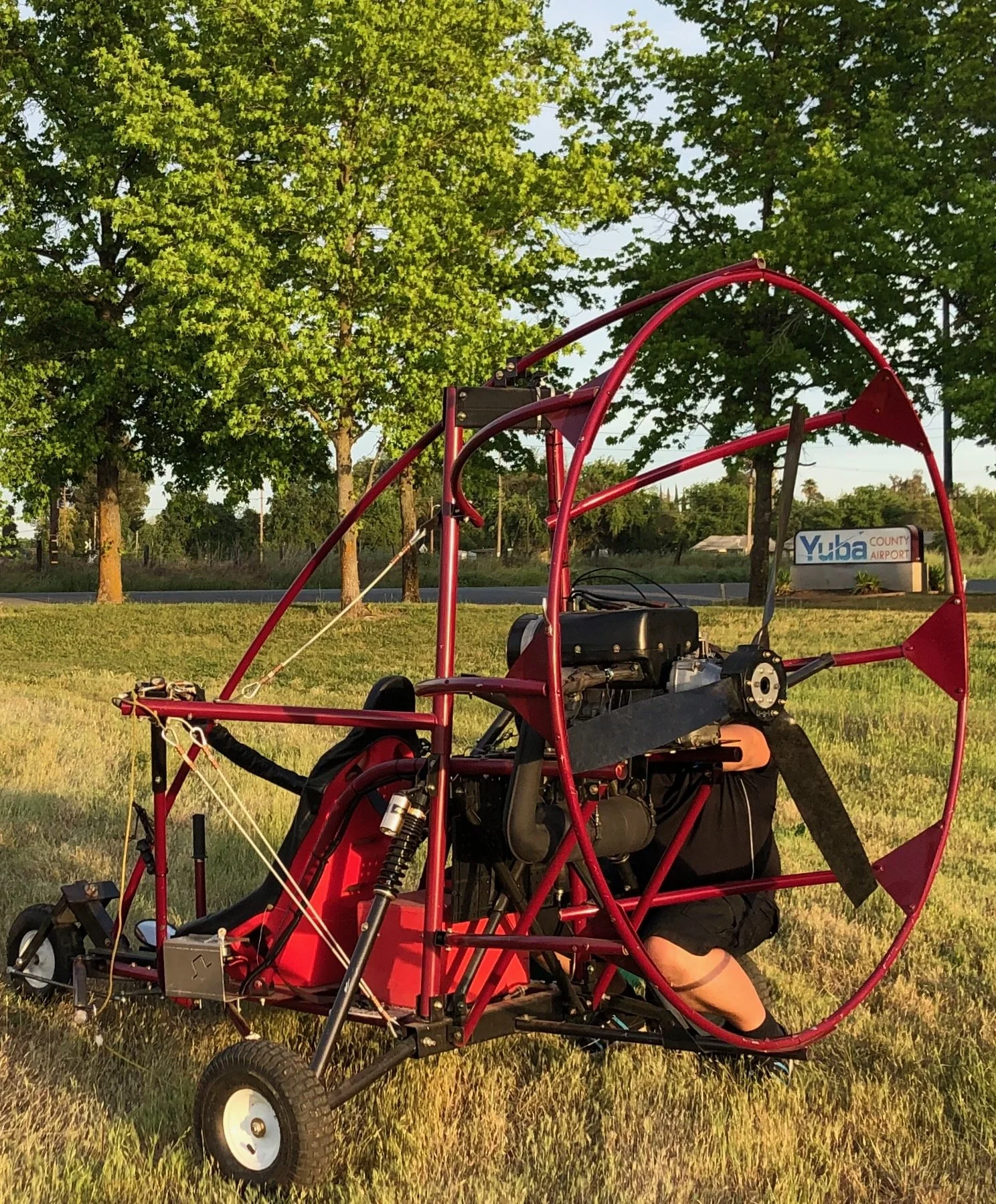 A man kneeling next to a powered paraglider with a red frame, black engine, and large circular propeller, on a grassy field with trees and a sign for Yuba County Airport in the background.