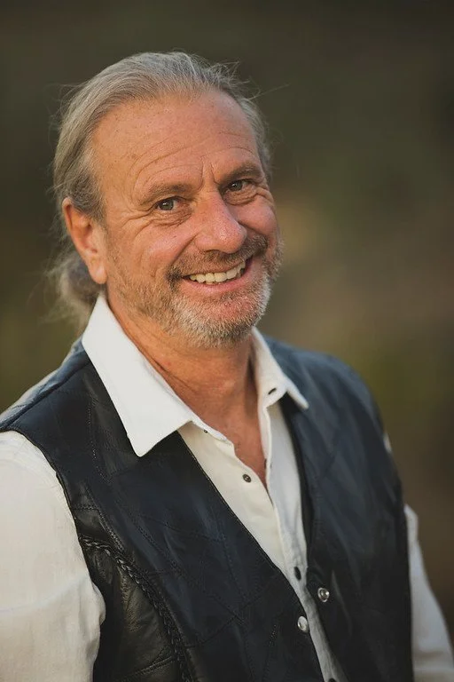 A smiling middle-aged man with gray hair and beard, wearing a white shirt and black leather vest, outdoors with a blurred background.