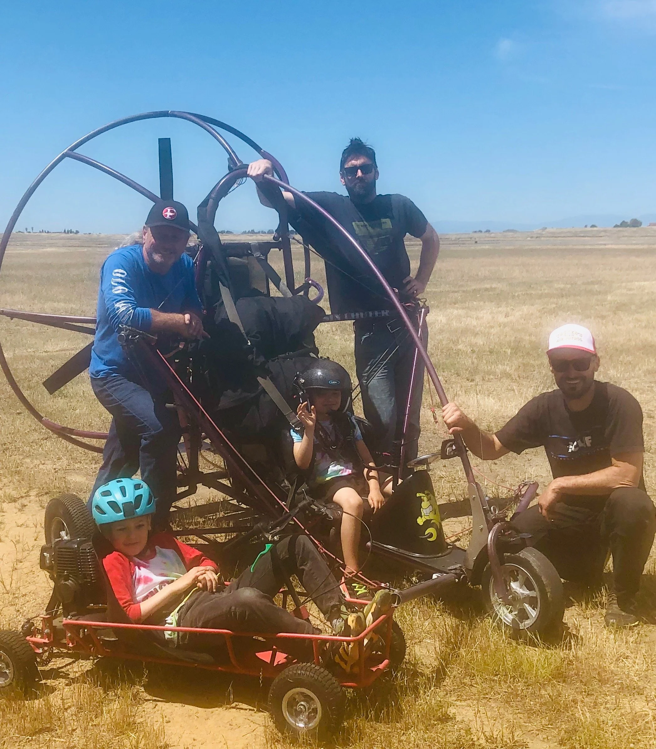 A group of five people, two children and three adults, are outdoors in a wide open field with clear skies, with some of them posing with a paramotor and a go-kart. Two children, one with a helmet, are sitting in the go-kart, while the adults are standing or kneeling beside them, one holding the go-kart's handle.
