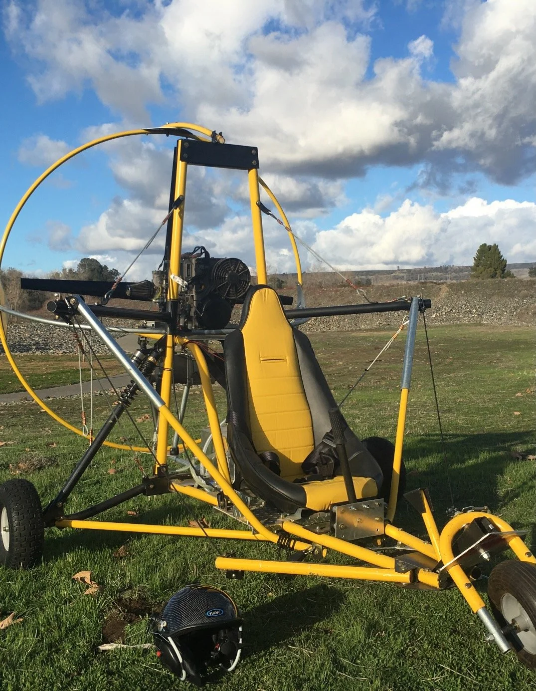 A yellow and black ultralight aircraft with a seat, engine, and frame, parked on grassy ground with a helmet nearby, under a partly cloudy sky.