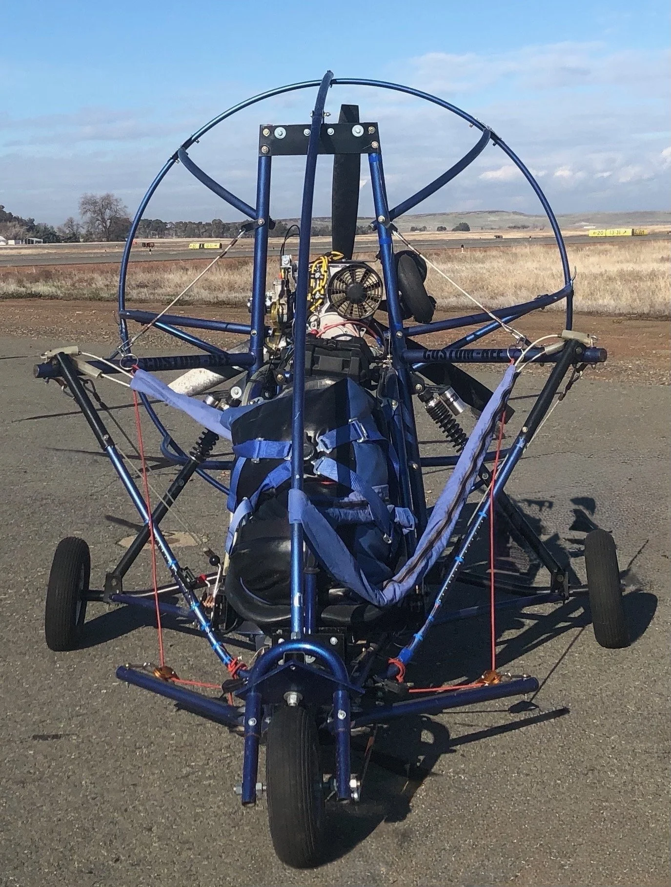 A blue ultralight aircraft with visible framework, open cockpit, and a pack placed inside, on a paved area in a rural landscape.