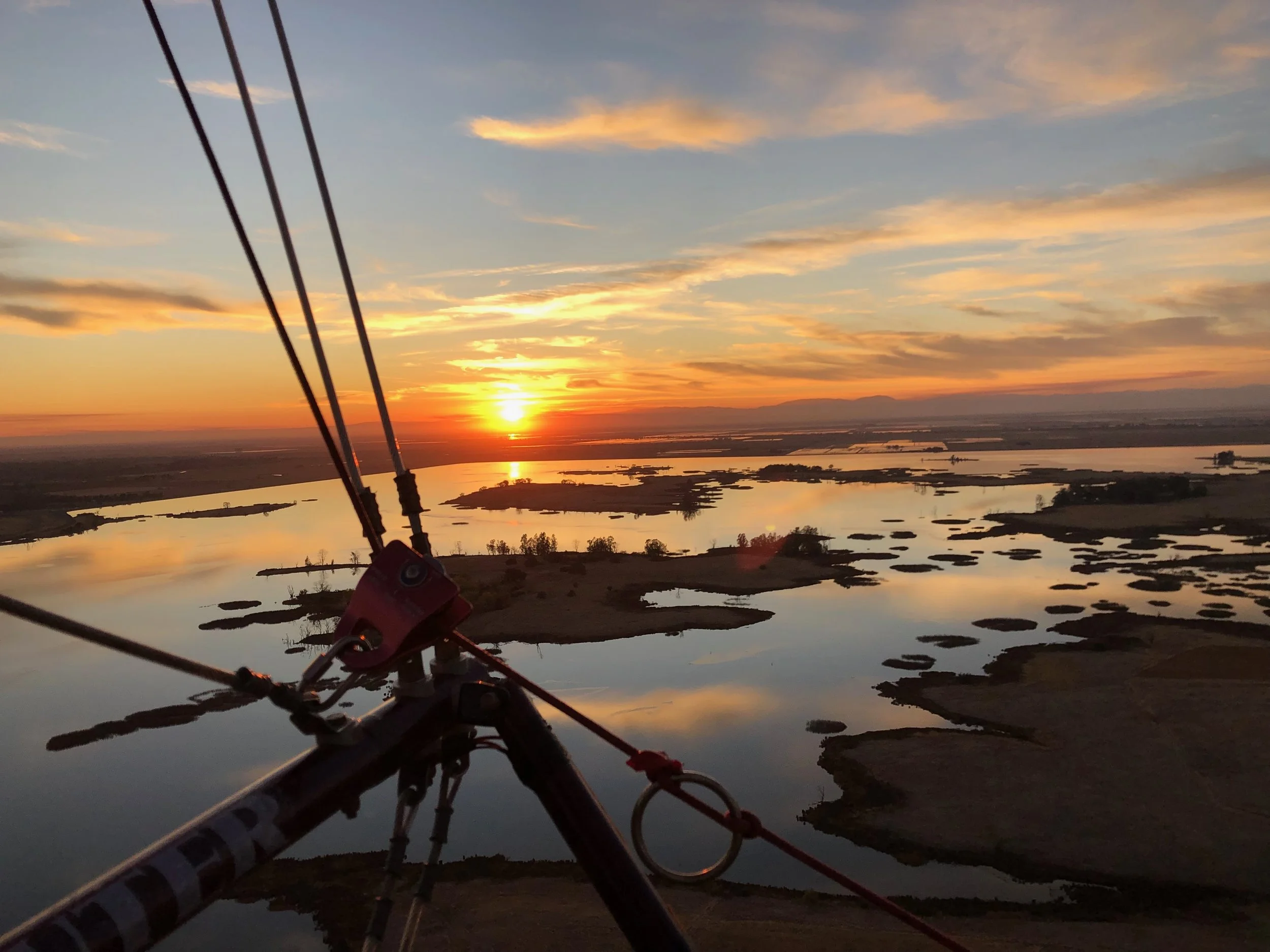 View from a hot air balloon at sunset, showing the basket's rigging, a river with multiple islands, and a colorful sky with clouds.