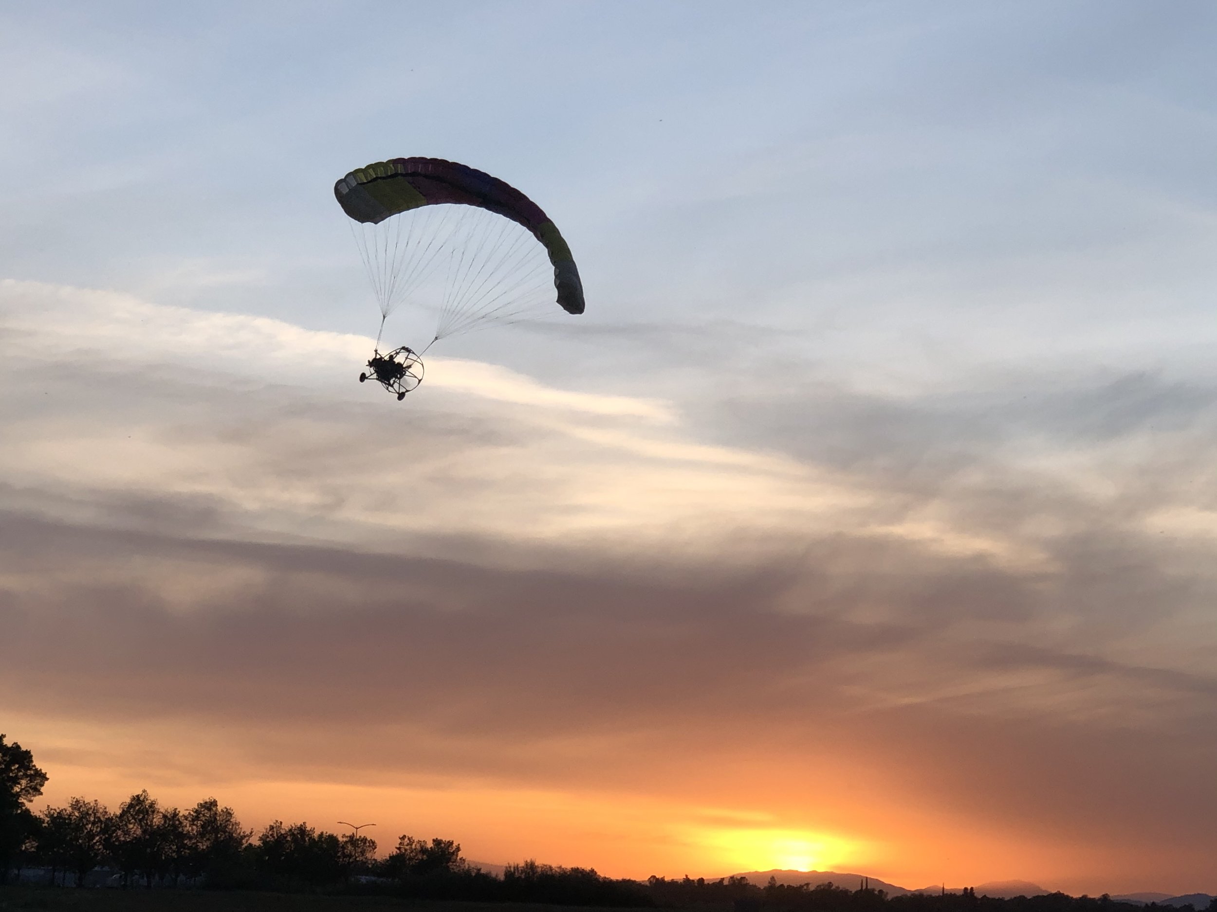A powered parachute flying in the sky during sunset with a colorful rainbow parachute, over a landscape with trees and mountains.
