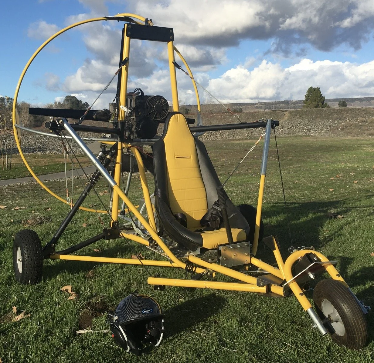 A yellow and black ultralight aircraft with a single seat and a propeller, parked on a grassy field, with a helmet placed on the ground nearby.