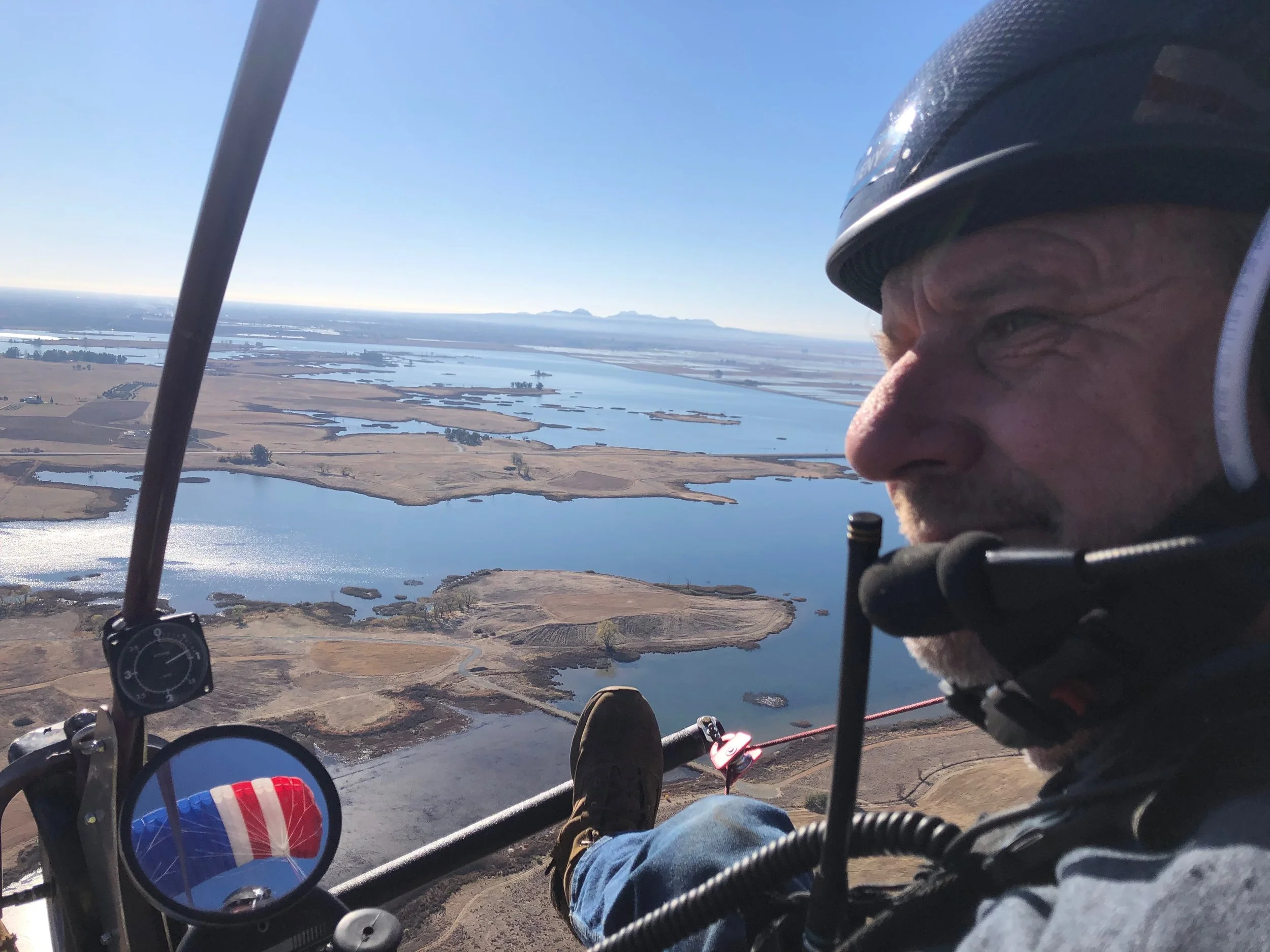 A man flying in a small aircraft over a body of water with land and islands, wearing a helmet and headset, with part of the aircraft's interior visible.