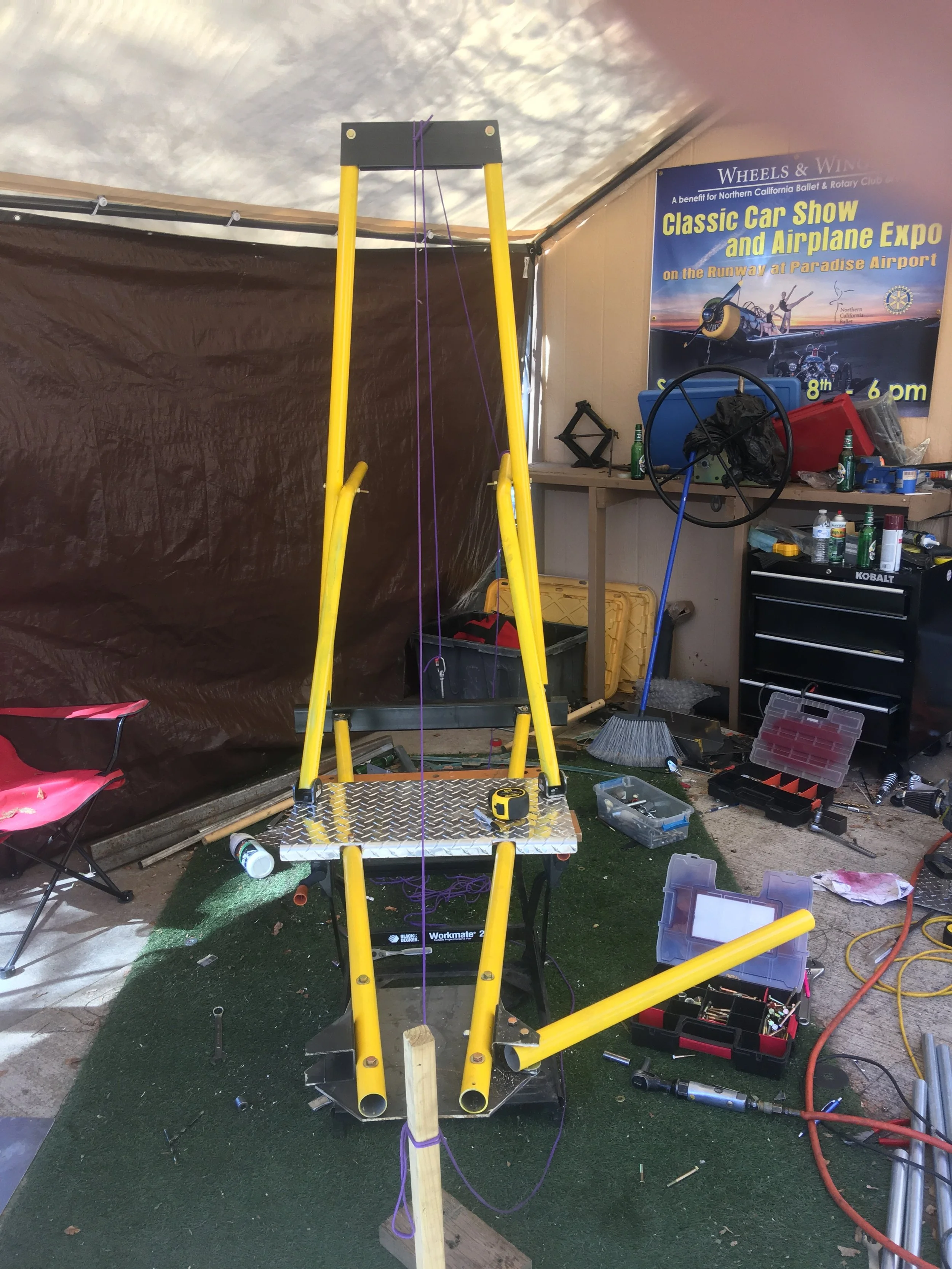 A work area with a yellow metal structure and a ladder, surrounded by tools and equipment, in front of a one-person flying contraption, with a poster advertising a classic car show and airplane expo in the background.