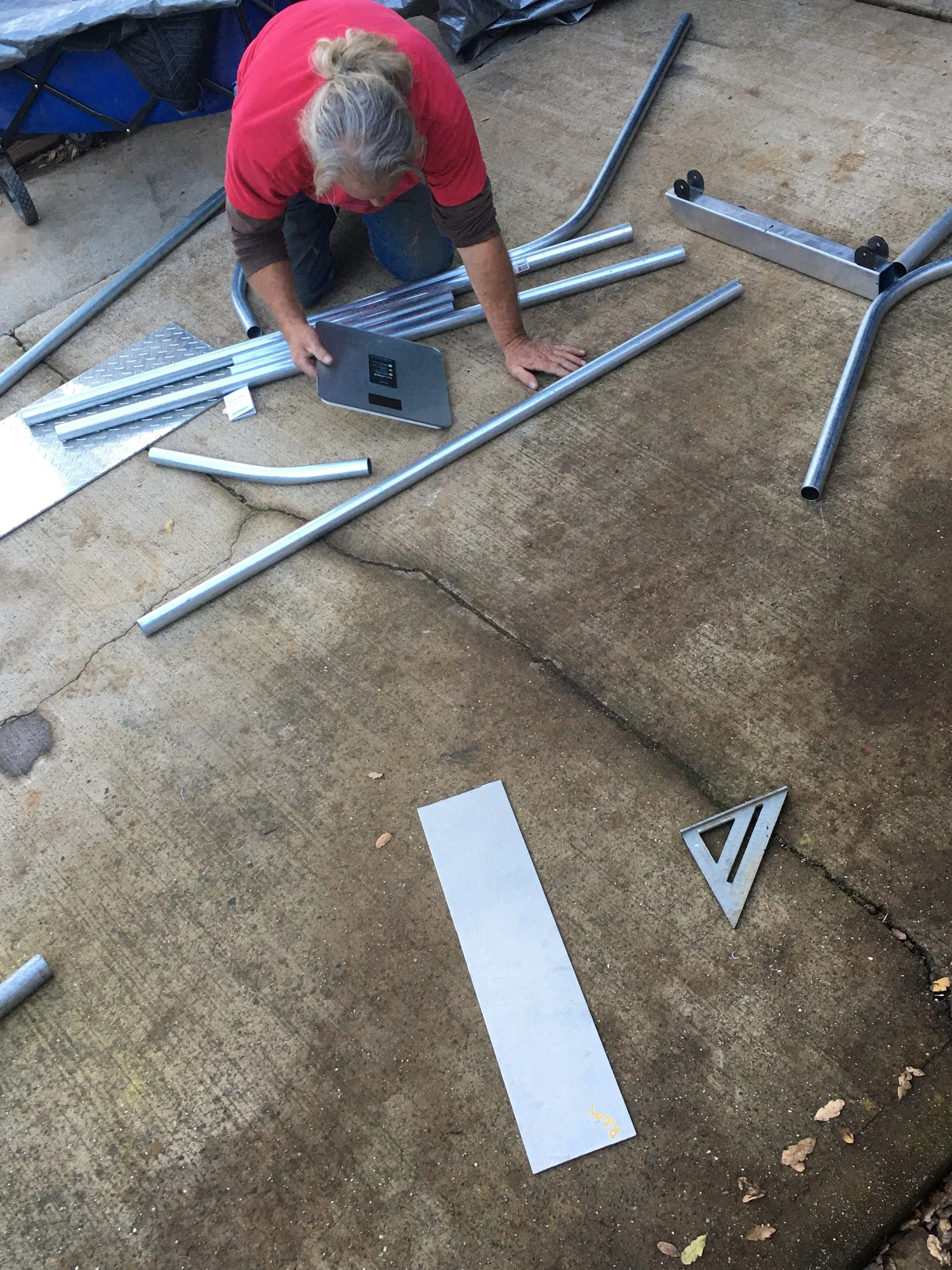 A person with long gray hair kneeling on a concrete surface, assembling metal pipes, with a digital scale or device nearby.