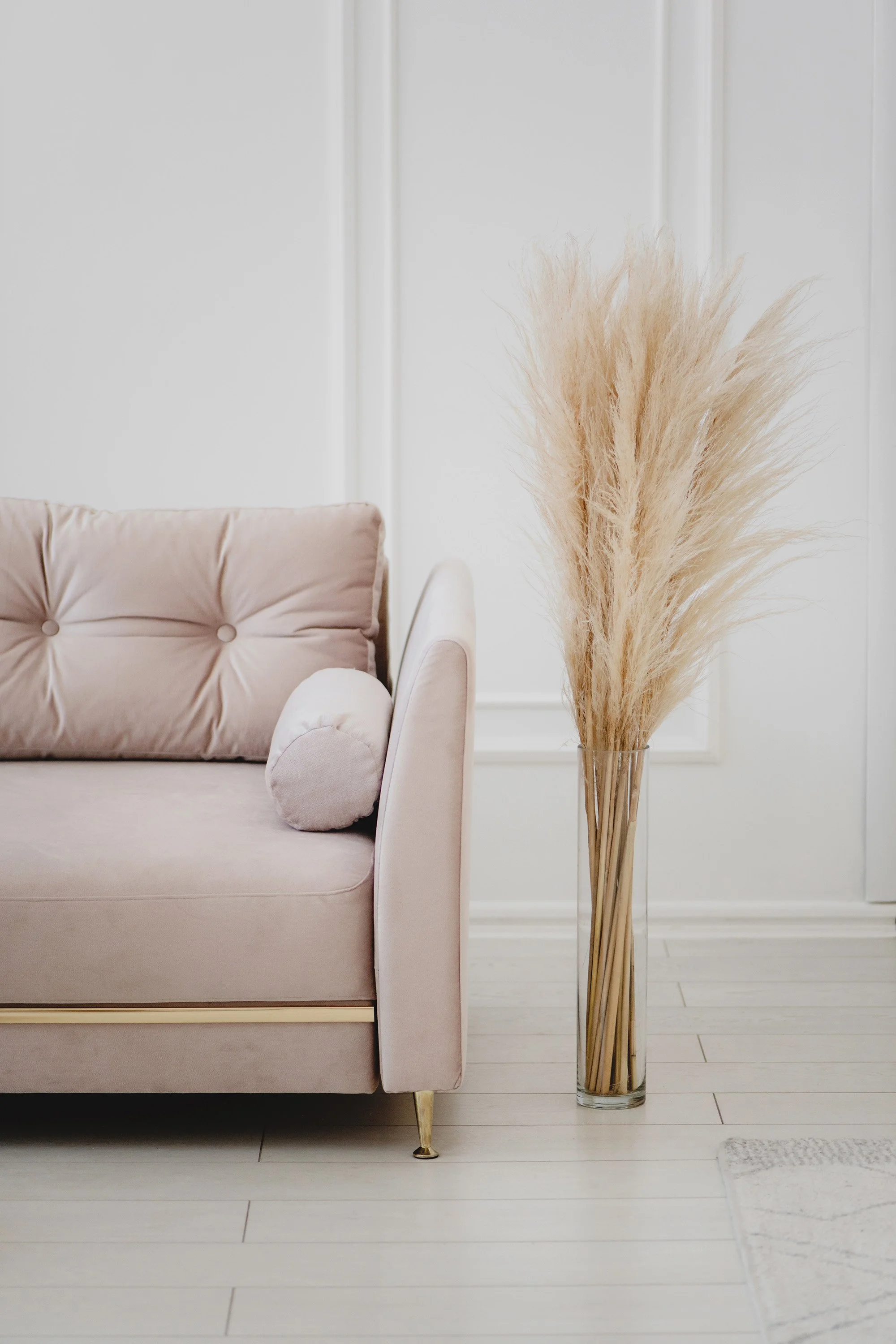 A closeup image of a blush, colored couch and next to it a tall vase on the floor with long wheat plants inside in front of a white wall