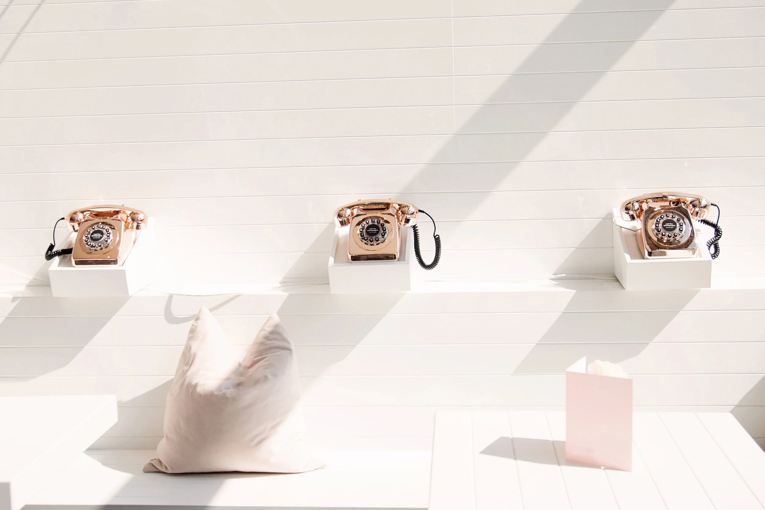 A wall that has a shelf with 3 old fashioned copper colored phones outside with the sunlight on them