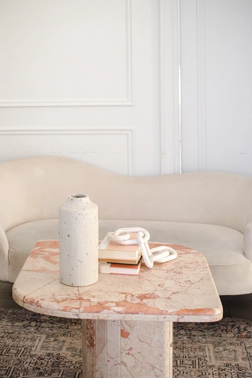 A closeup shot of a coffee table with an empty ceramic vase and a few books next to it stacked below on a big chain decoration with a beige couch in the background against a white wall with panelling