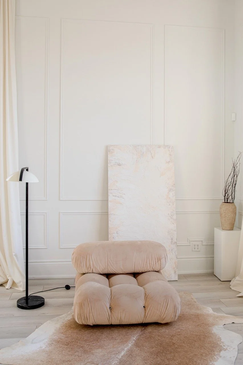 A living area with a single beige couch on the floor on top of a fluffy rug, a long and tall painting up against the wall with panelling, a standing lamp next to it and a vase on a long rectangle base