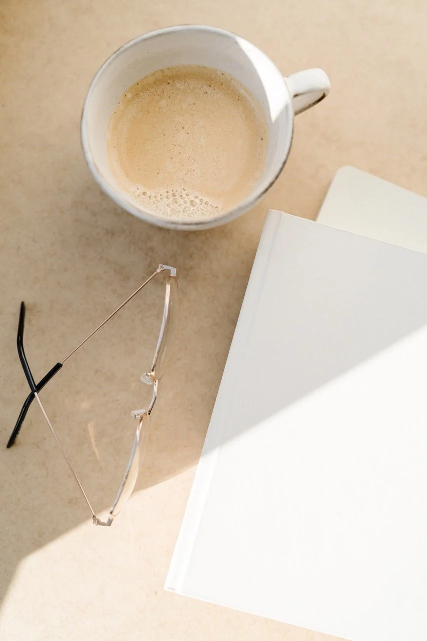 A closeup flatlay of a cream colored table that has a pair of reading glasses, a glass of half full coffee and a closed, white hard cover journal