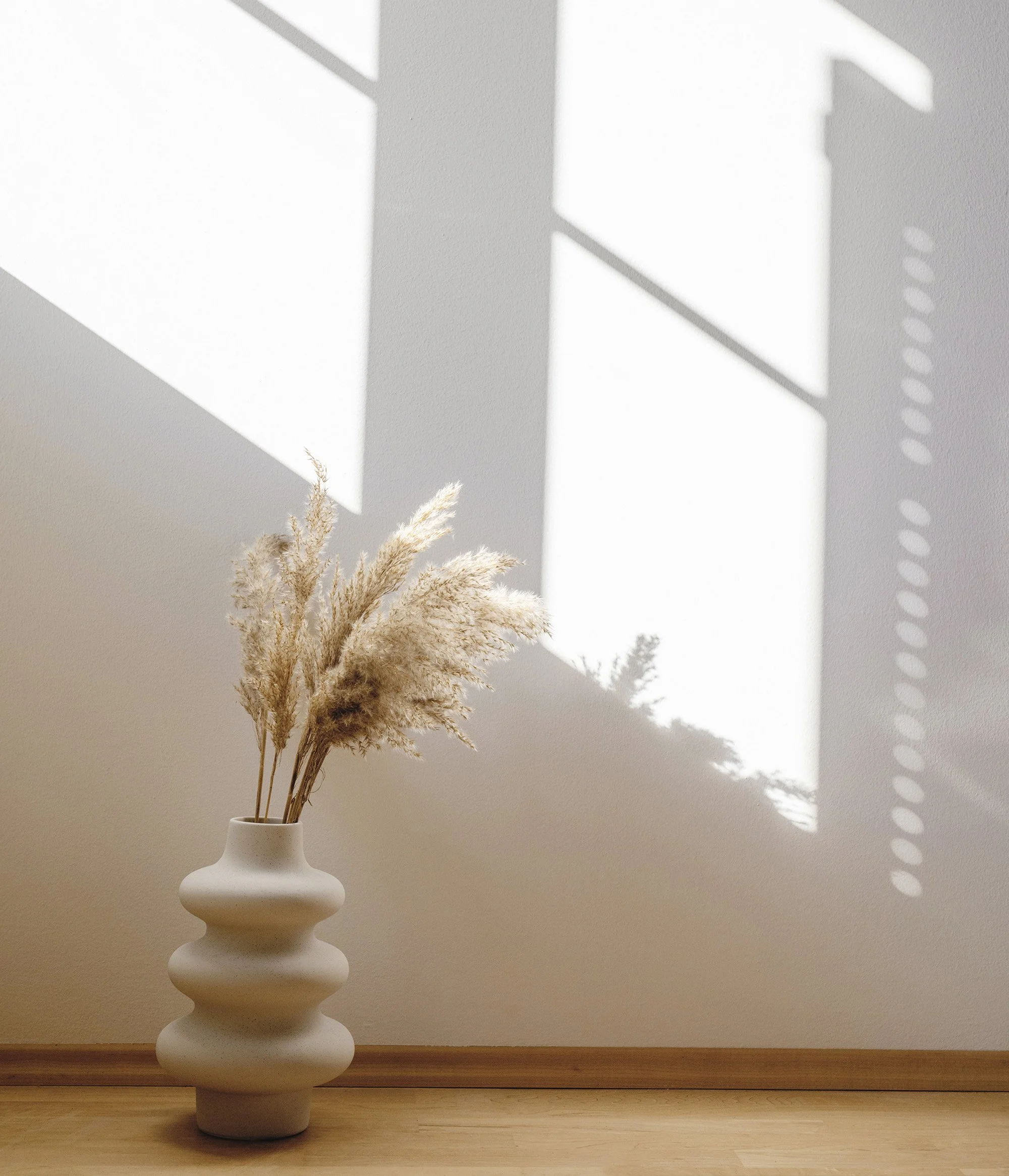 a ceramic vase on the wooden floor next to a window that has the sunlight showing on the wall casting shadows of the window