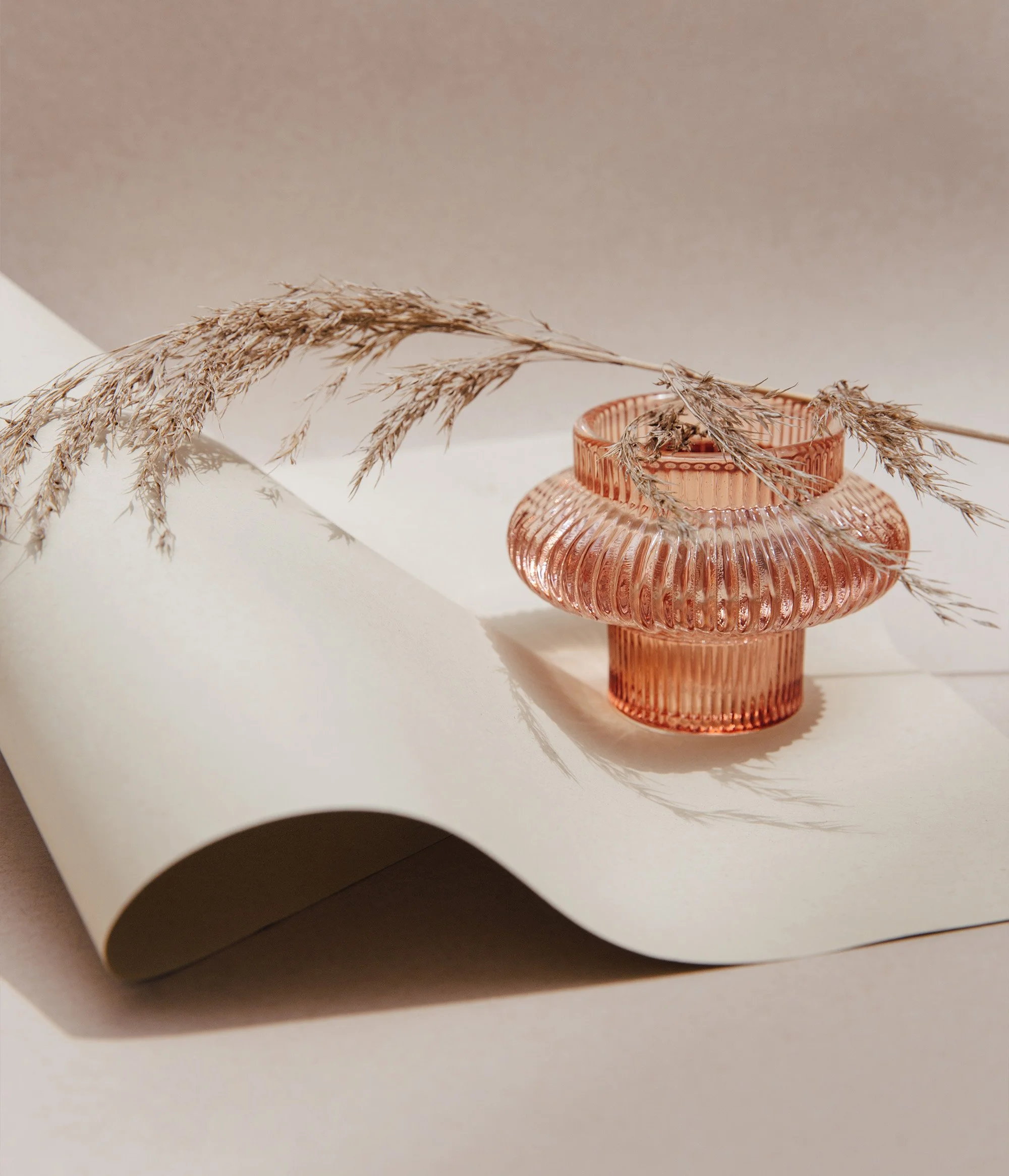 a rose gold wide glass vase on top of a rolled poster board on the floor with a wheat plant laying on top of the vase