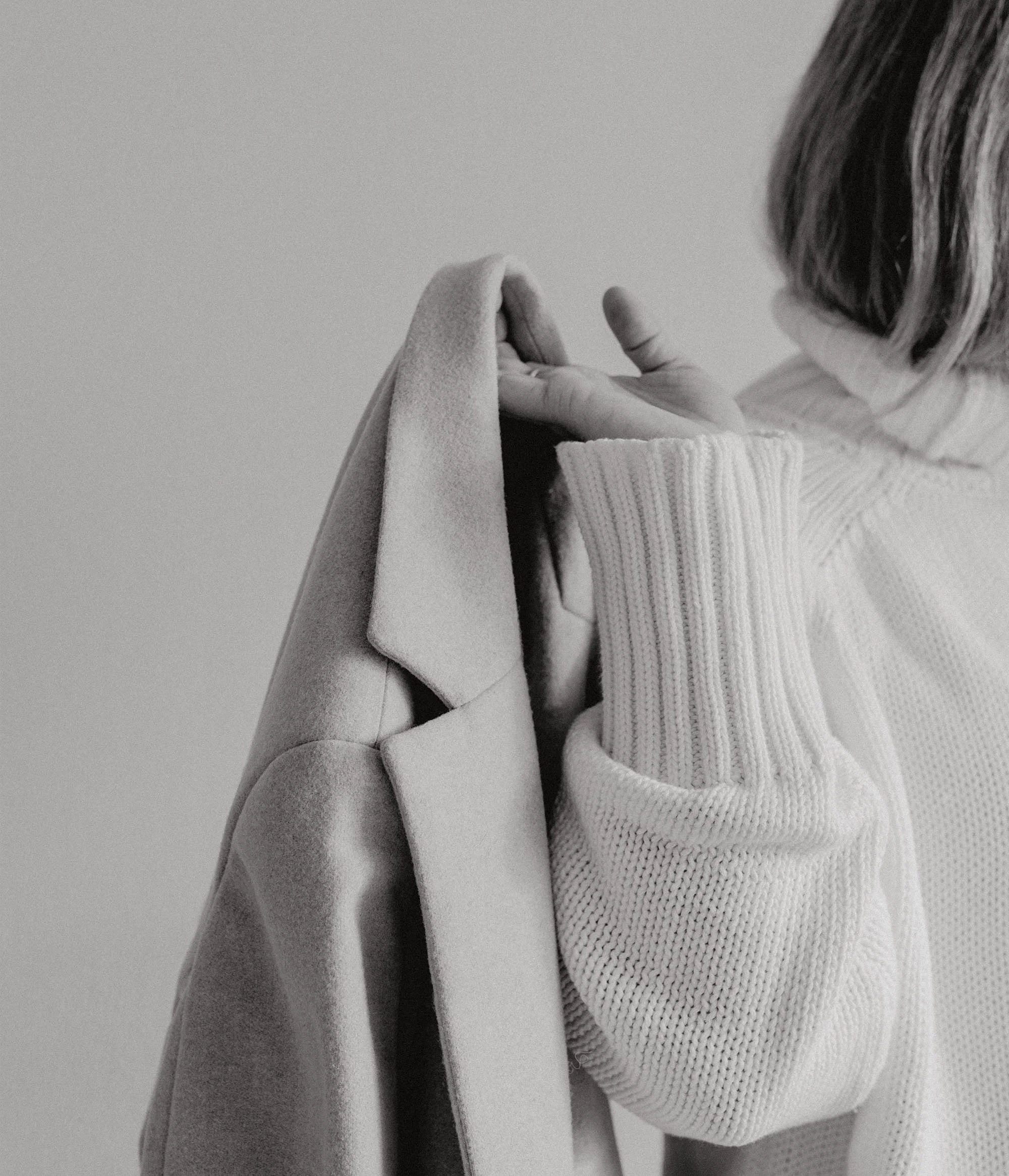 a black and white image of a woman hanging her coat from her hand that is next to her shoulder while wearing a white sweater