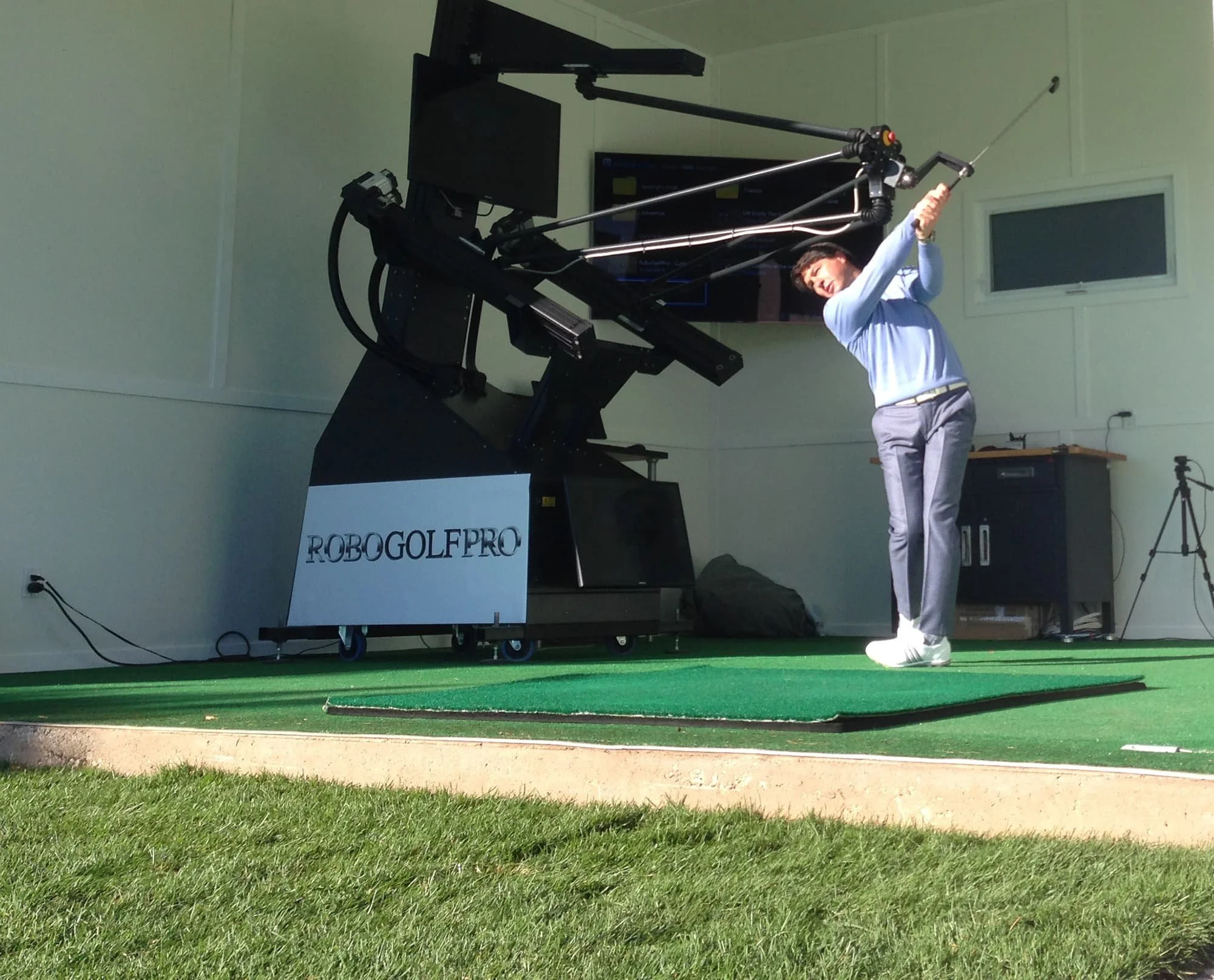 A woman practicing golf indoors with a robotic golf simulator, labeled 'ROBOGOLF PRO,' positioned on a green artificial turf.