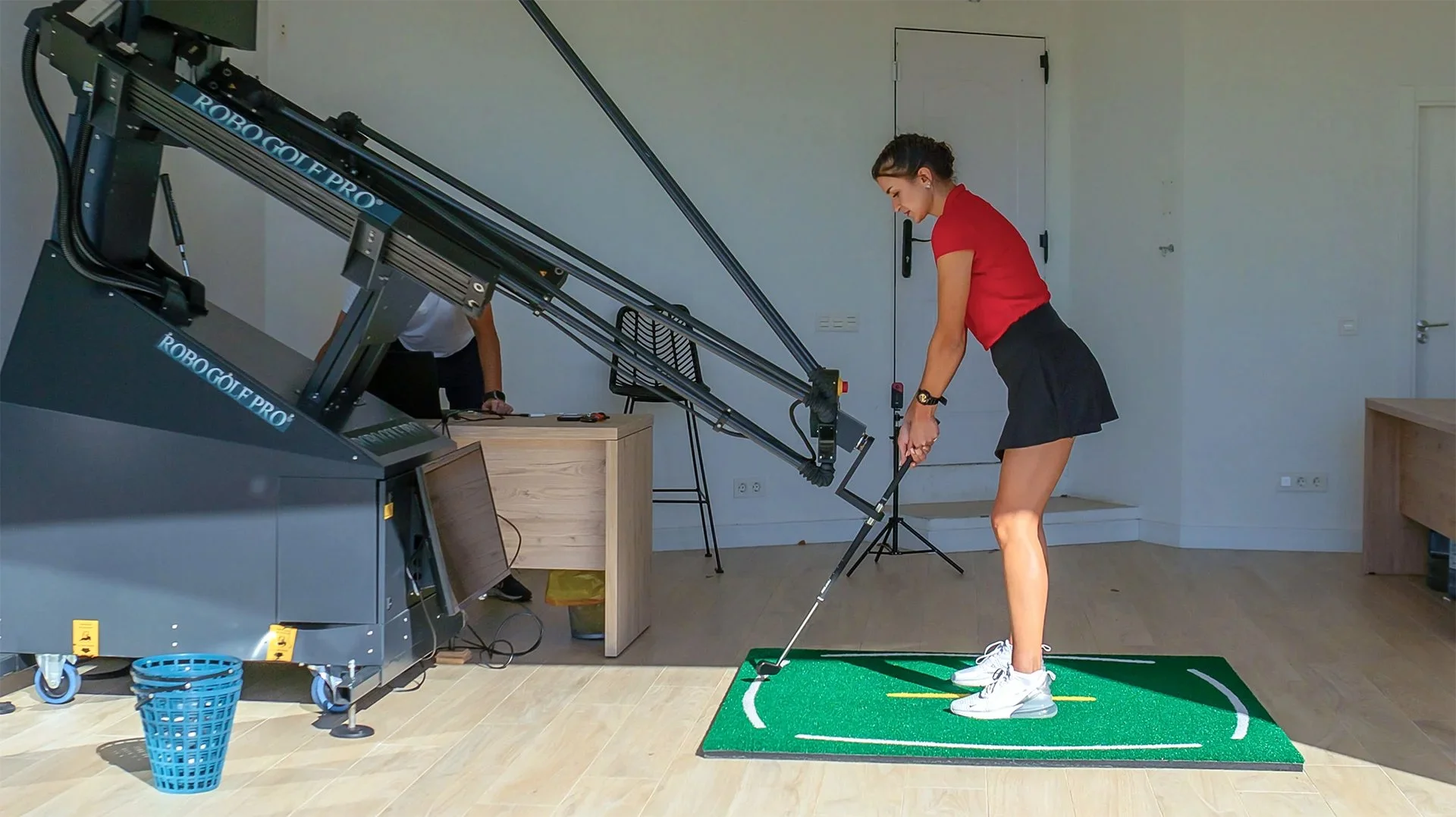 Woman practicing golf indoors on a green mat with a golf club, a large robotic golf swing training machine nearby, and a person working on a desk in background.