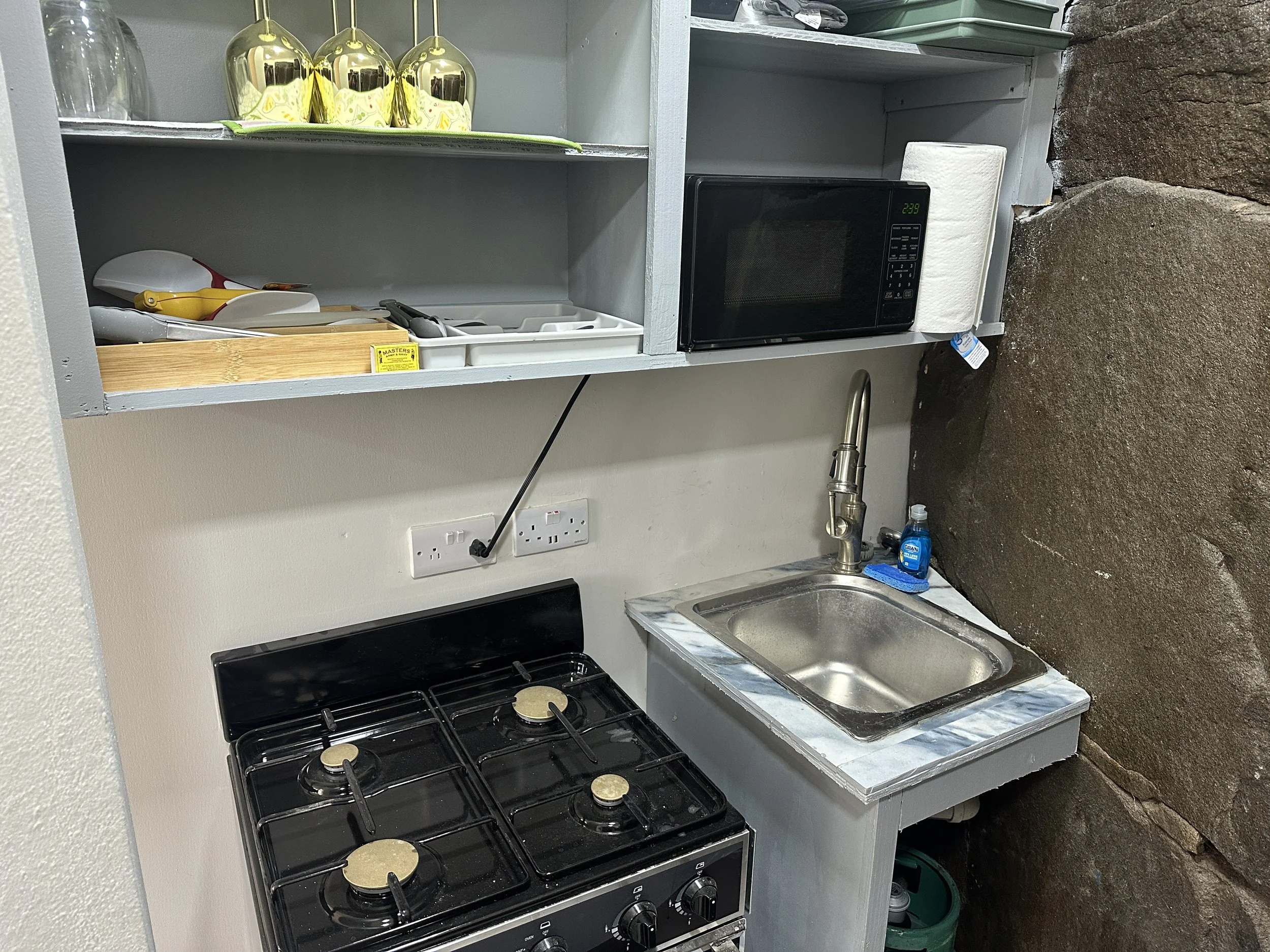 Compact kitchen area with a gas stove, small sink, microwave, paper towel roll, and shelves with kitchen utensils, glassware, and miscellaneous items.