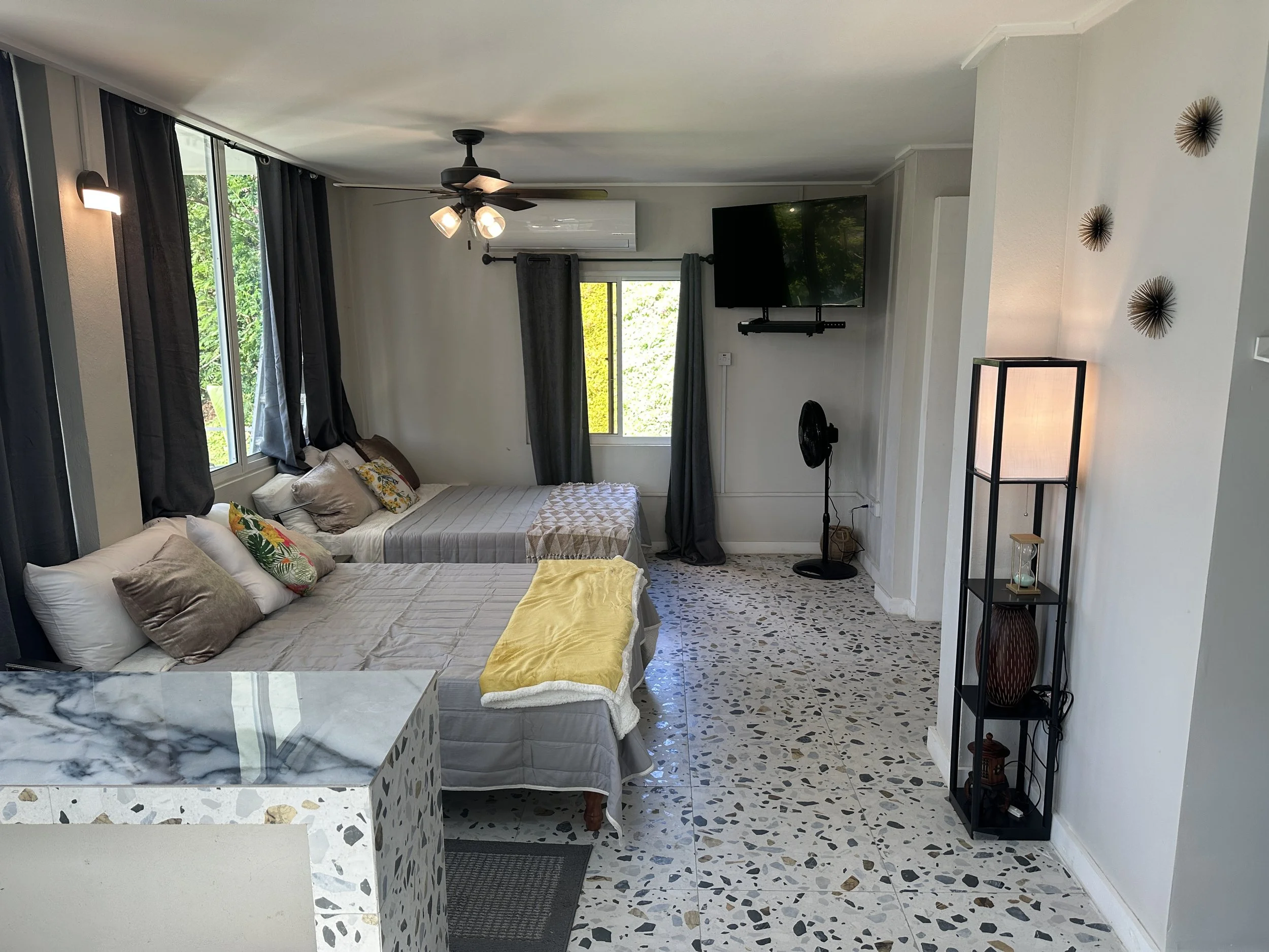 Living room with two beds, dark curtains, a wall-mounted TV, a standing fan, a tall floor lamp, and decorative wall art.