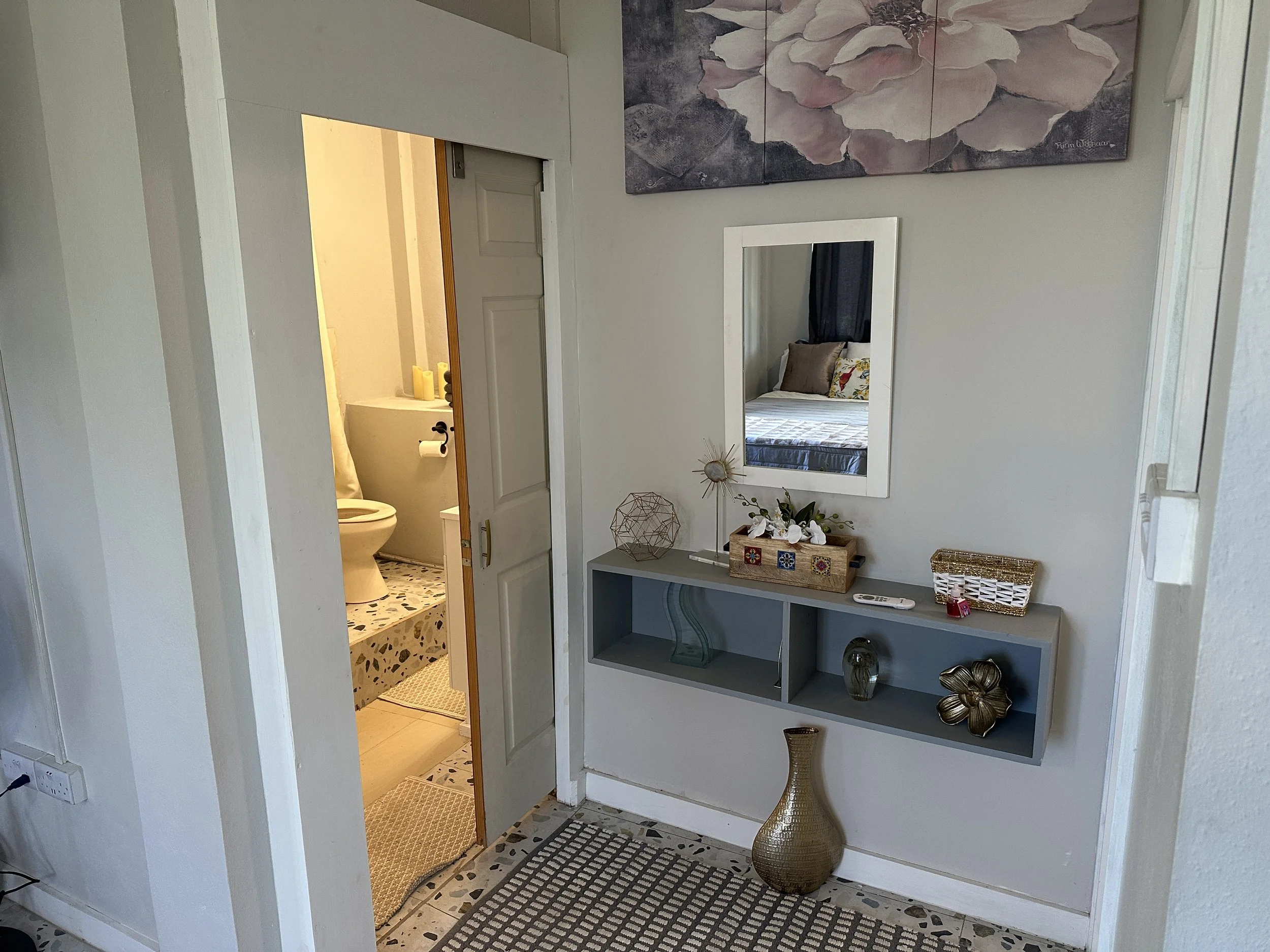 View of a small hallway with a mirror on the wall, a shelf with decorative items, and a door leading to a bathroom with a toilet and bathtub visible.