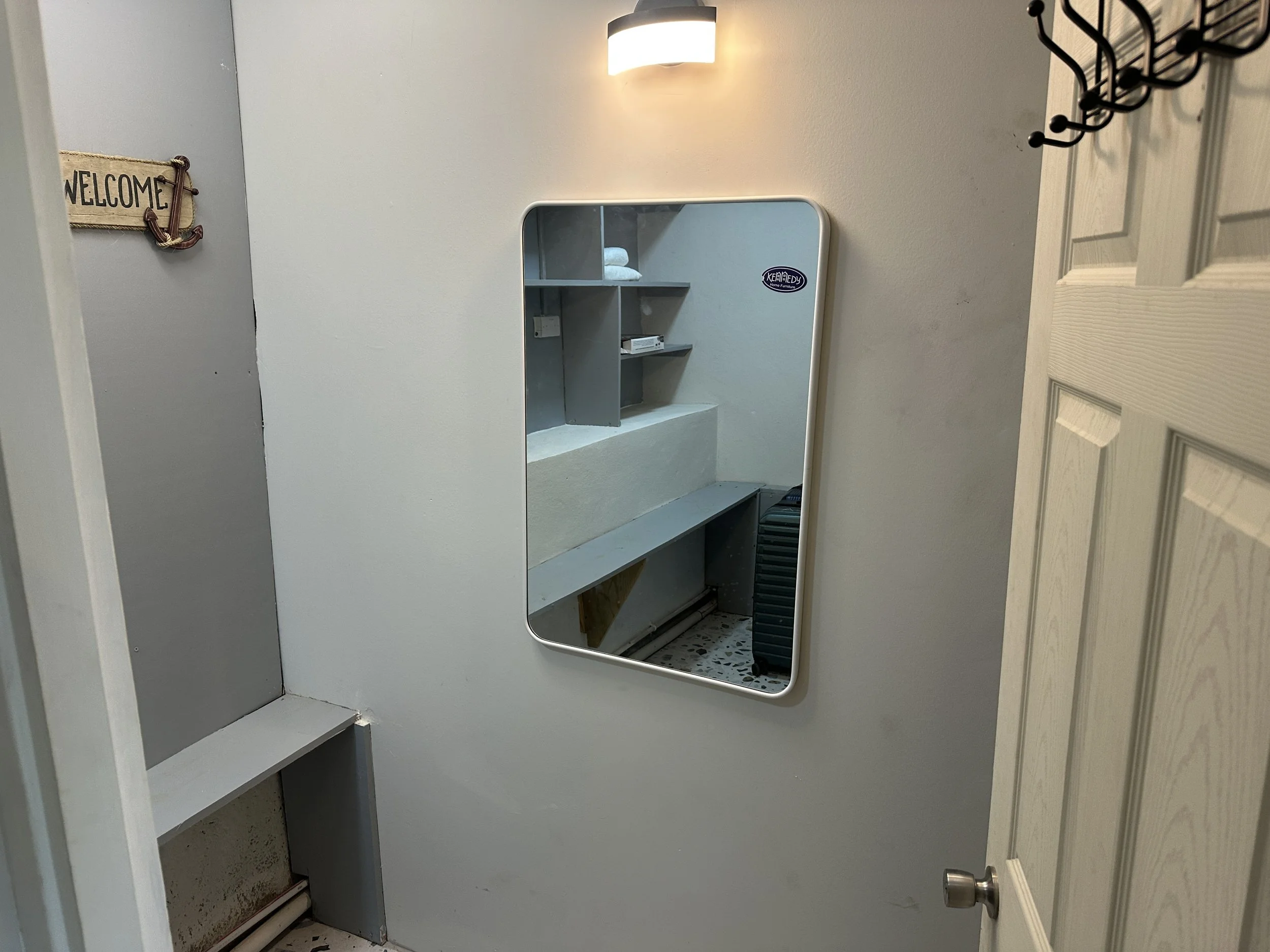 A small room with a mirror, a desk, and wall shelving, with a welcome sign on the wall, and a door partially open.