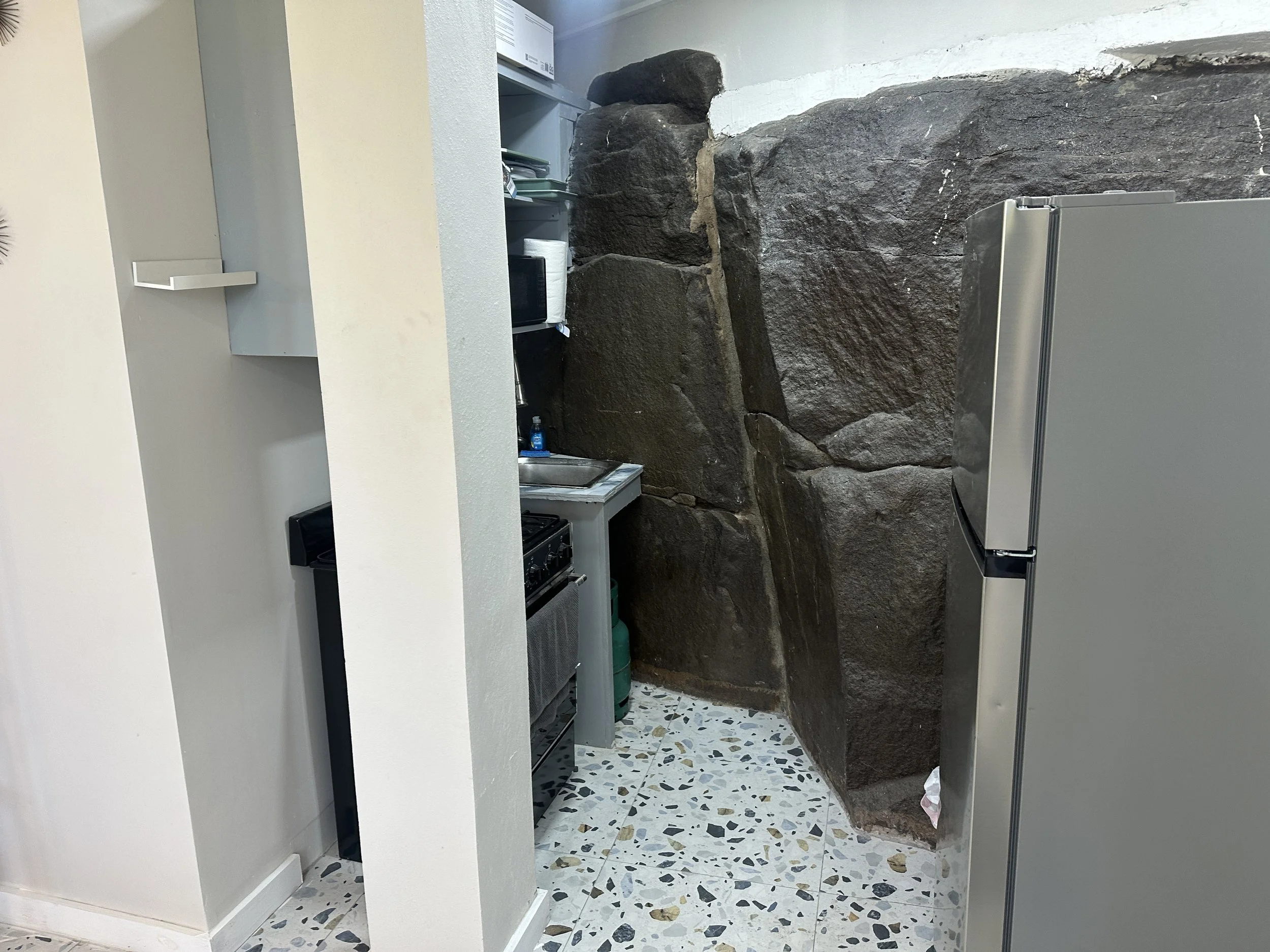 A small kitchen with a stone wall and terrazzo flooring, containing a refrigerator, a stove, a sink, and a shelf with paper towels and other items.