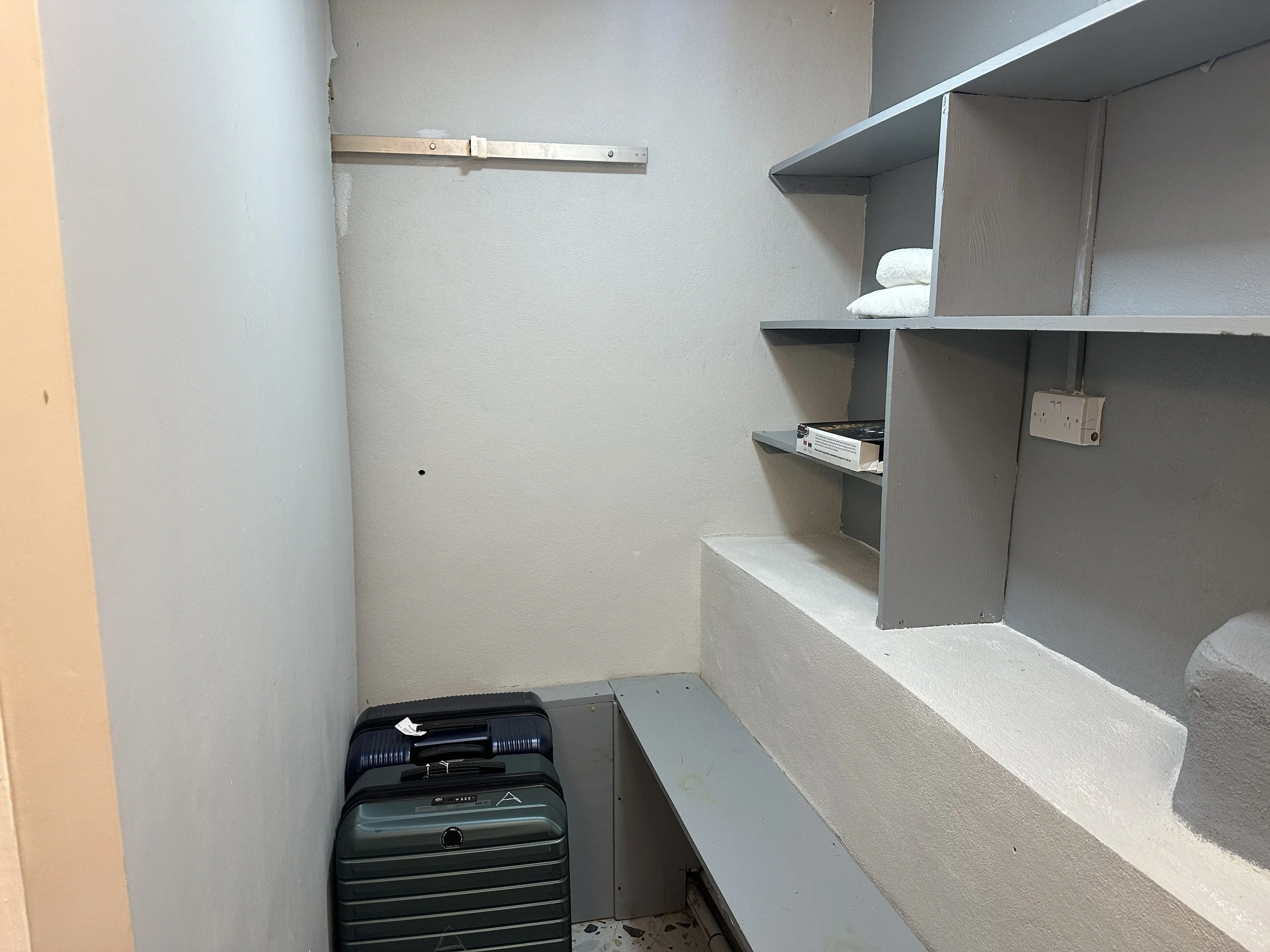 A small, mostly empty storage room or closet with a light gray wall, gray wooden shelves, a shelf with folded white towels, a box, a white electrical outlet, and two suitcases on the floor.