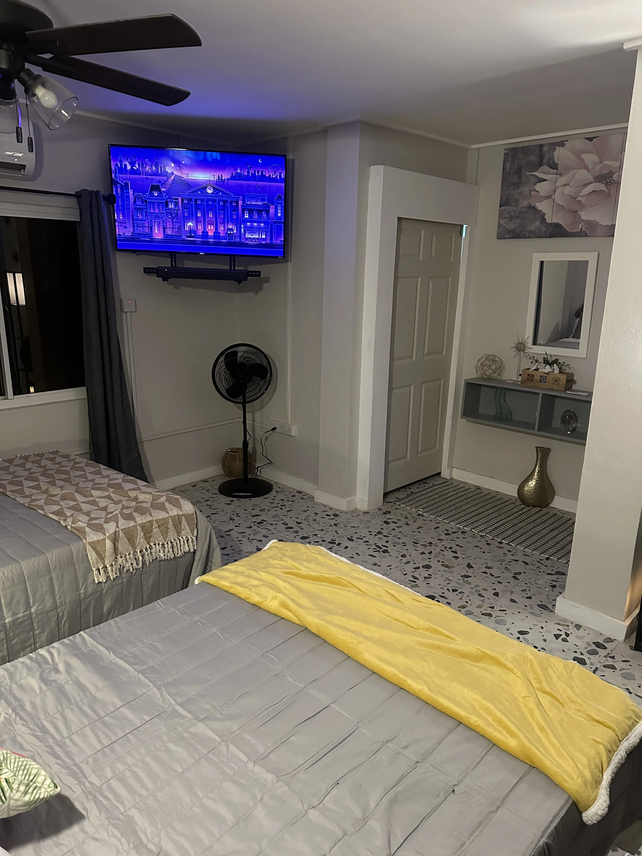 This is a cozy living room with two beds, a wall-mounted TV, a standing fan, a window with dark curtains, and decorative items on shelves and a side table.