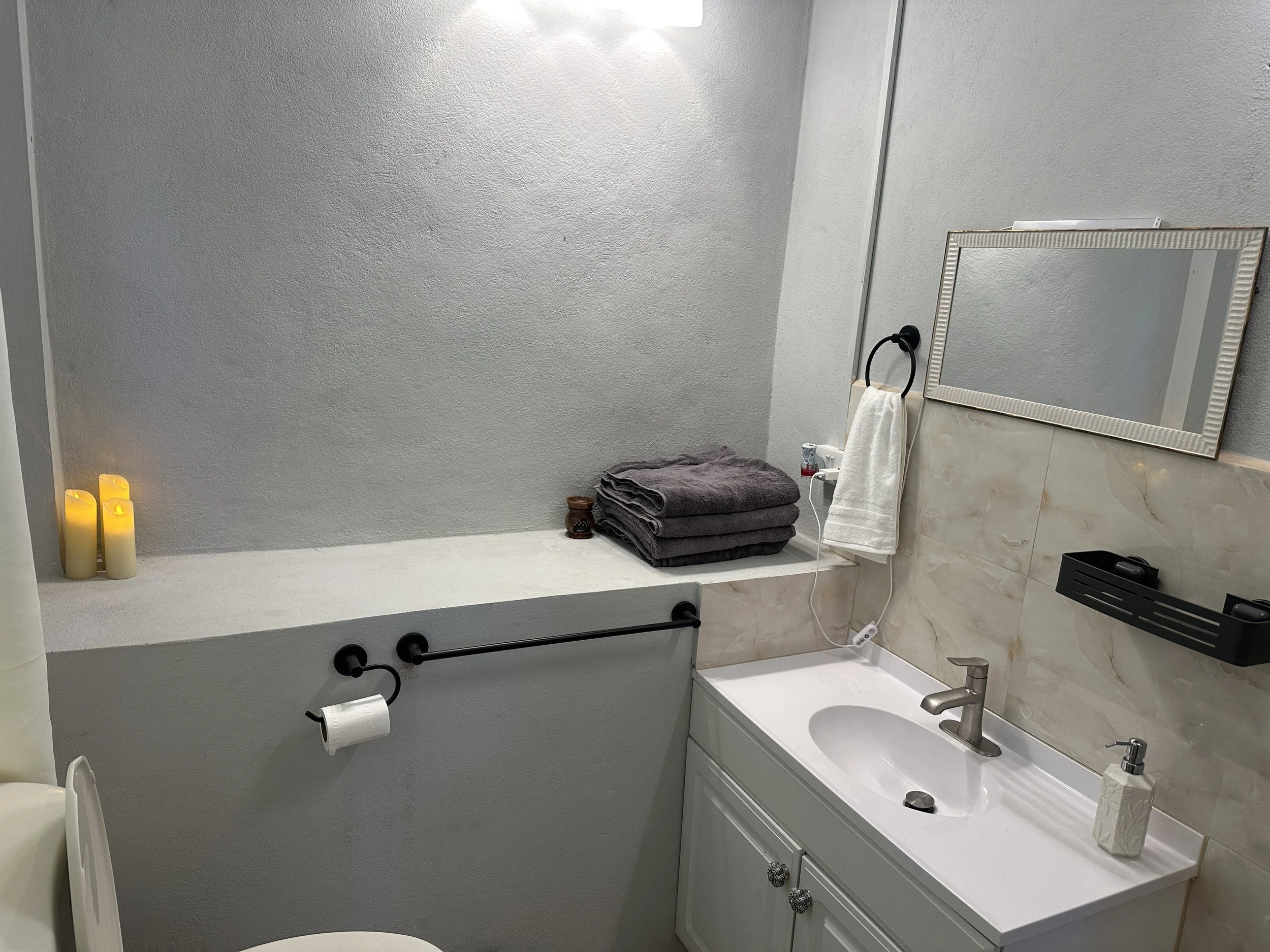 Bathroom with gray and white decor, candles on a ledge, folded towels on a counter, mirror, wall-mounted hand towel ring, black dispenser, and a small sink with a soap dispenser.