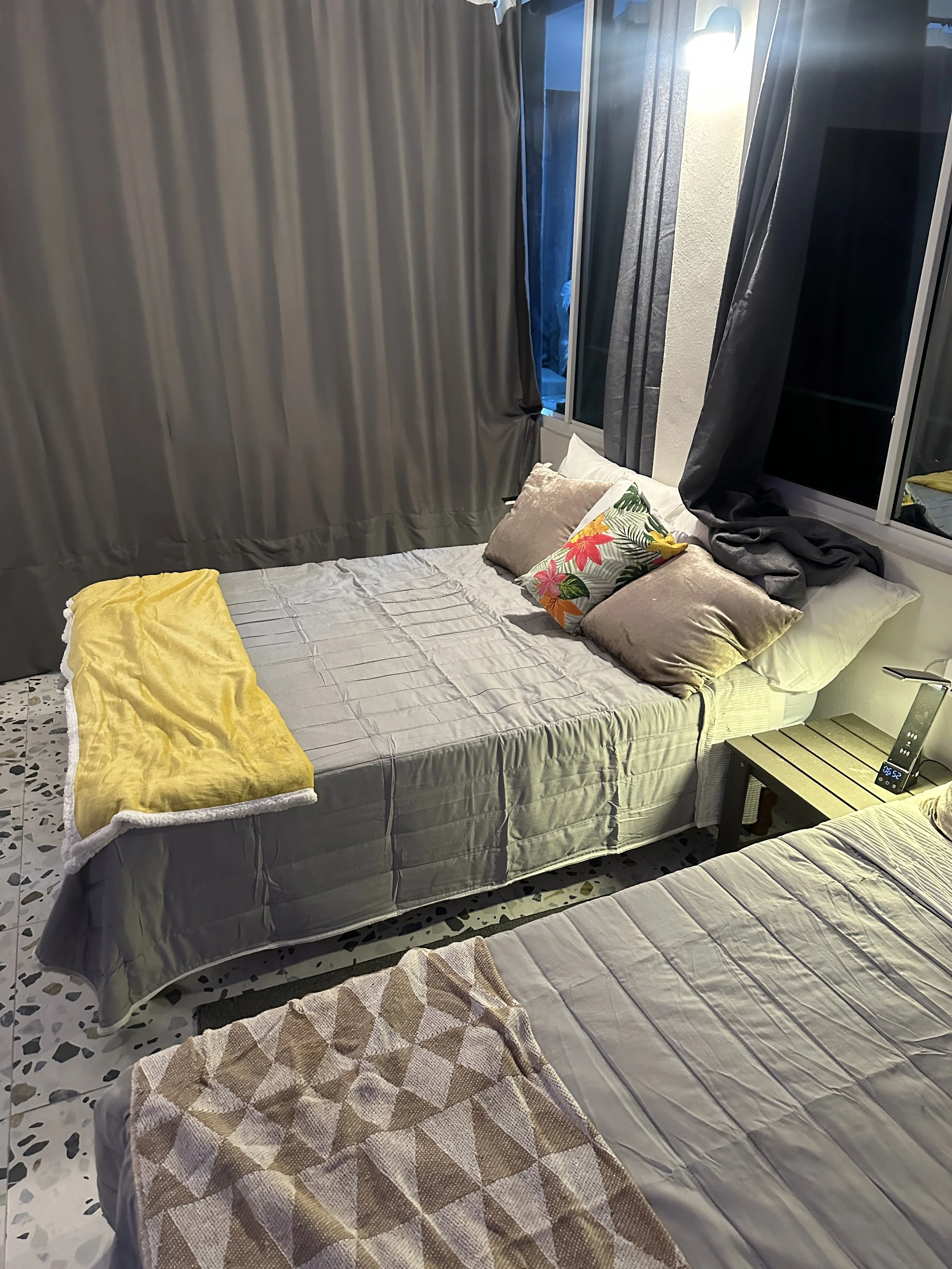 Two beds in a bedroom with beige bedspreads, multiple pillows, a nightstand with a clock, and curtains covering the windows.