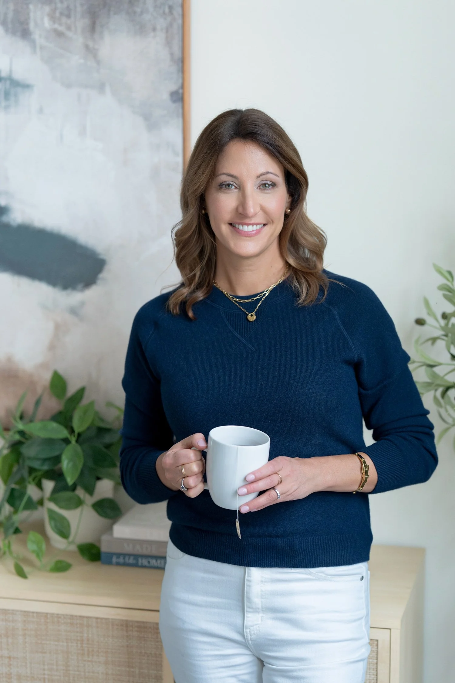 A woman smiling and holding a white mug, standing indoors near a light-colored wall and some greenery. Jenn Goonan Portrait